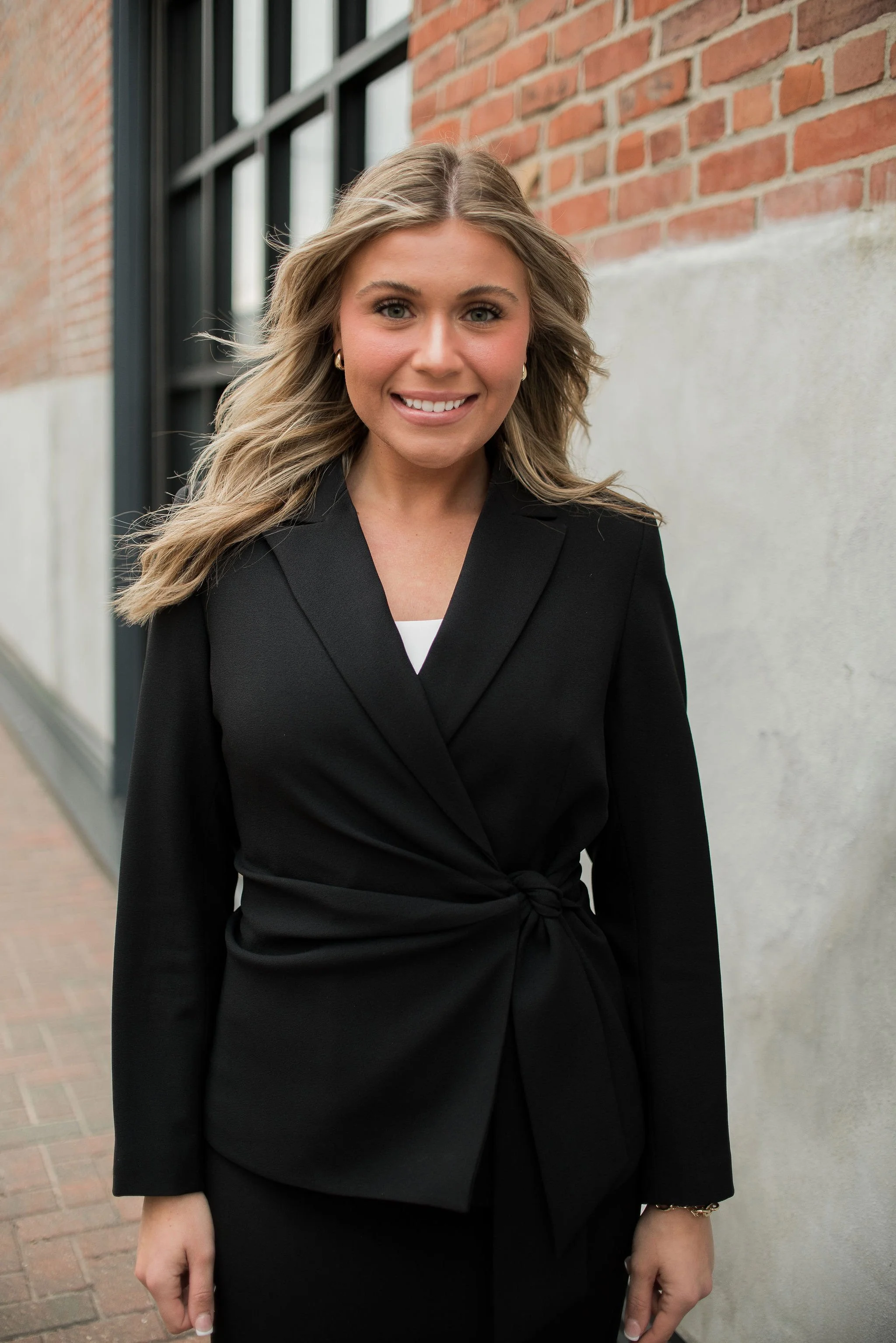 A woman with blonde, wavy hair smiling at the camera, wearing a black blazer with a tied waist, standing outside near a brick wall and window.