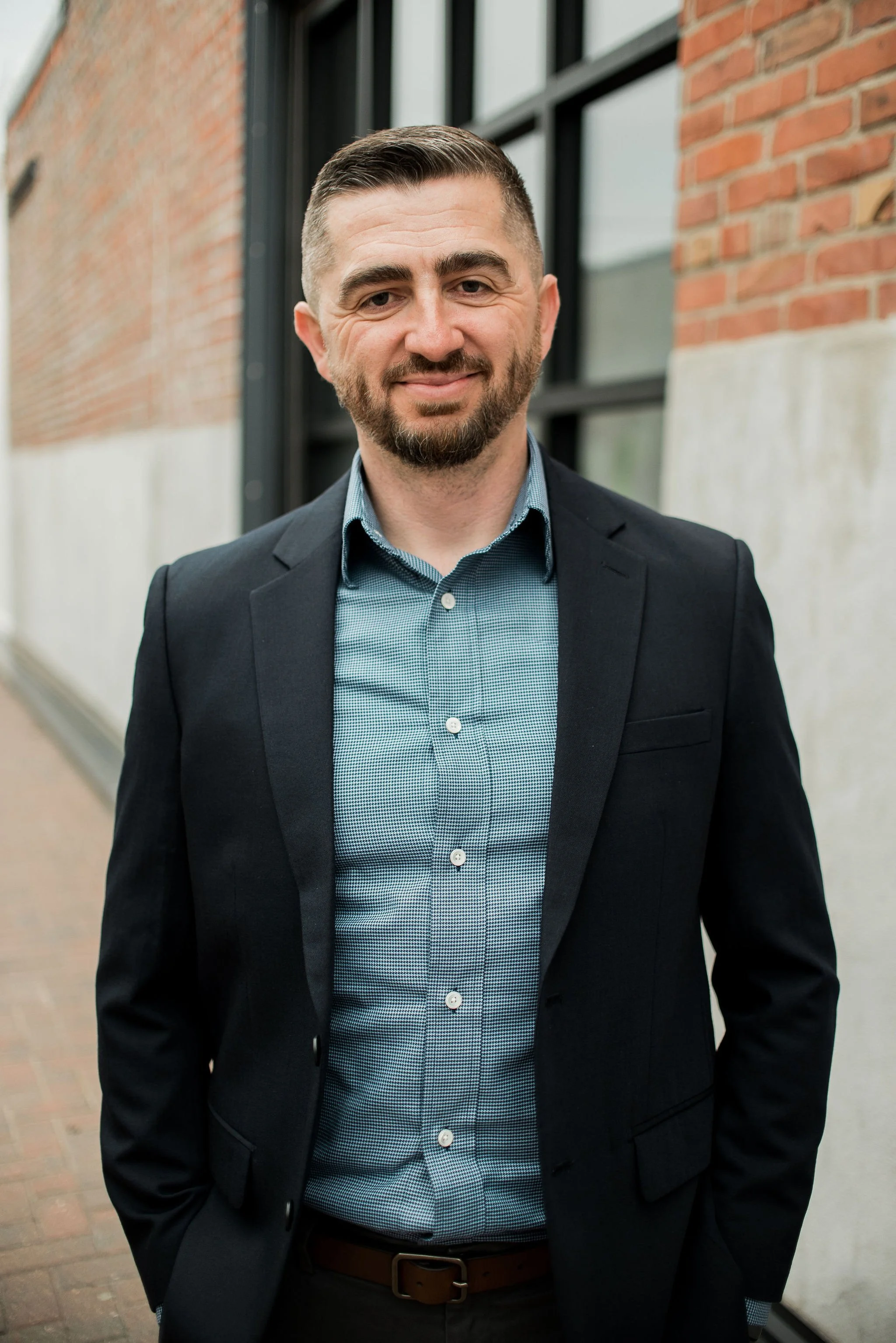 A man in a business suit outdoors, standing near a brick and concrete wall with large windows, smiling at the camera.