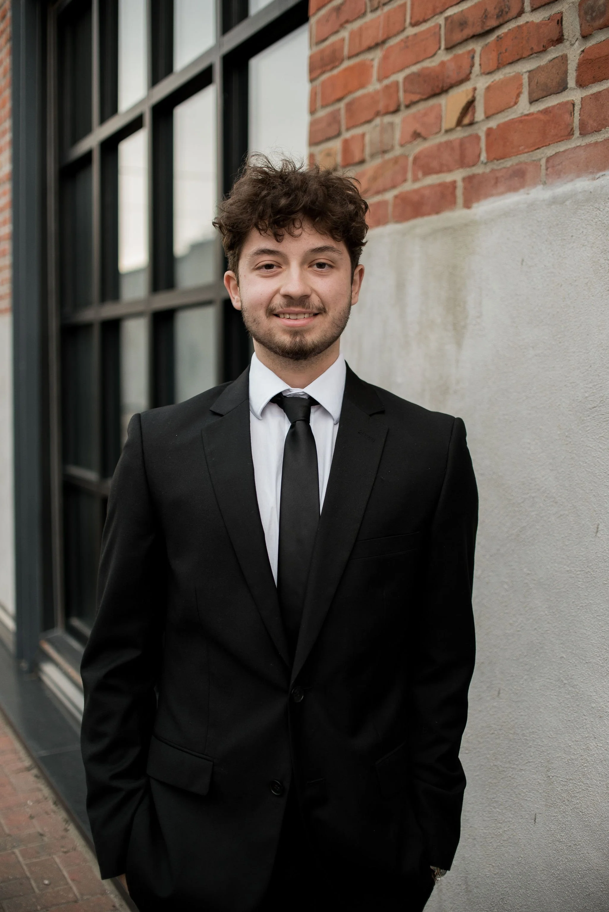A young man with curly brown hair, wearing a black suit, white shirt, and black tie, standing outdoors in front of a brick and white wall, smiling at the camera.