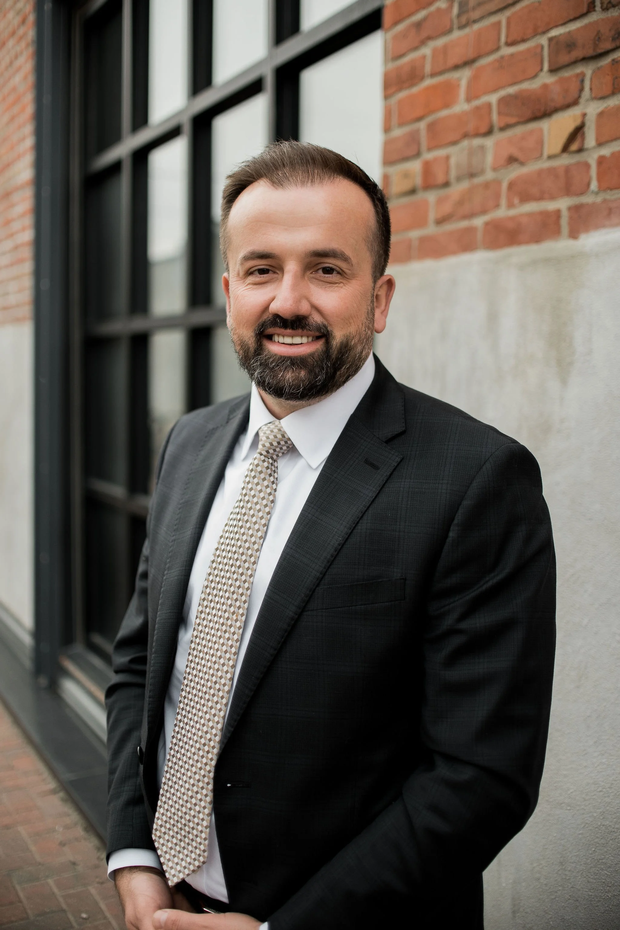 A man in a suit smiling, standing outdoors against a brick and concrete wall.