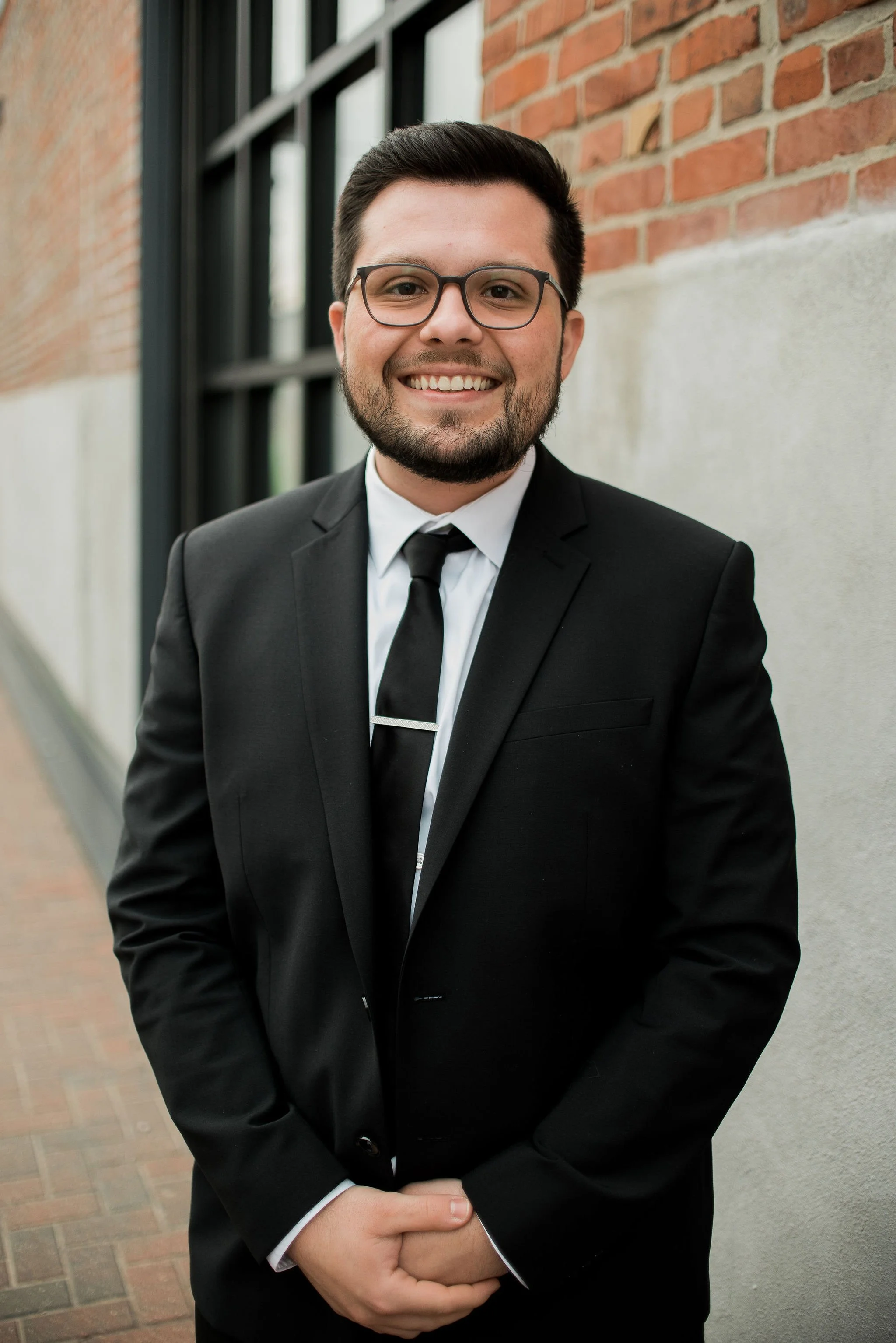 A man with dark hair, glasses, and a beard, wearing a black suit and tie, smiling and standing outdoors against a brick and concrete wall.