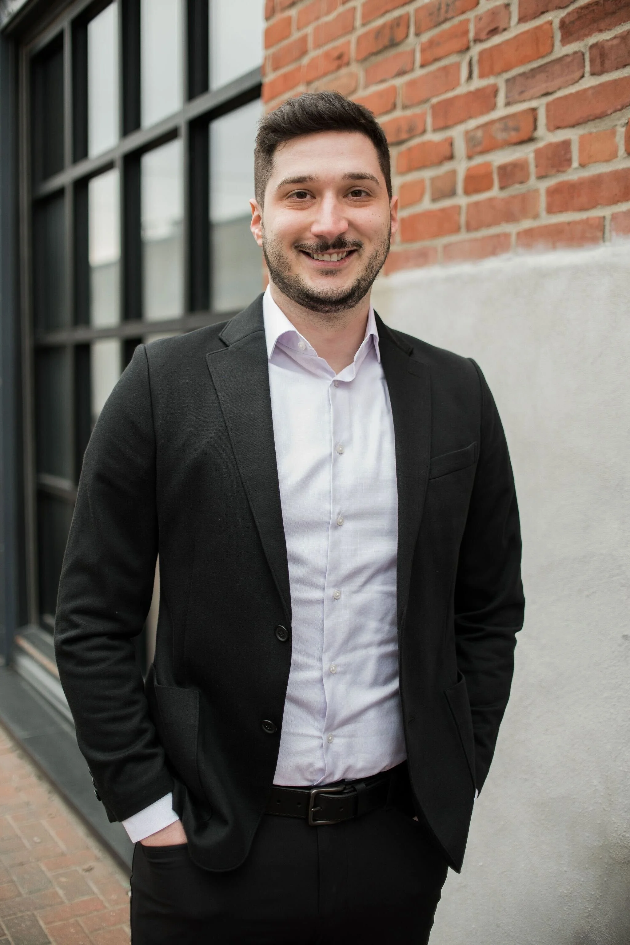 A young man in a black suit and white shirt standing outdoors near a brick and concrete wall, smiling at the camera. Mirza Alibasic