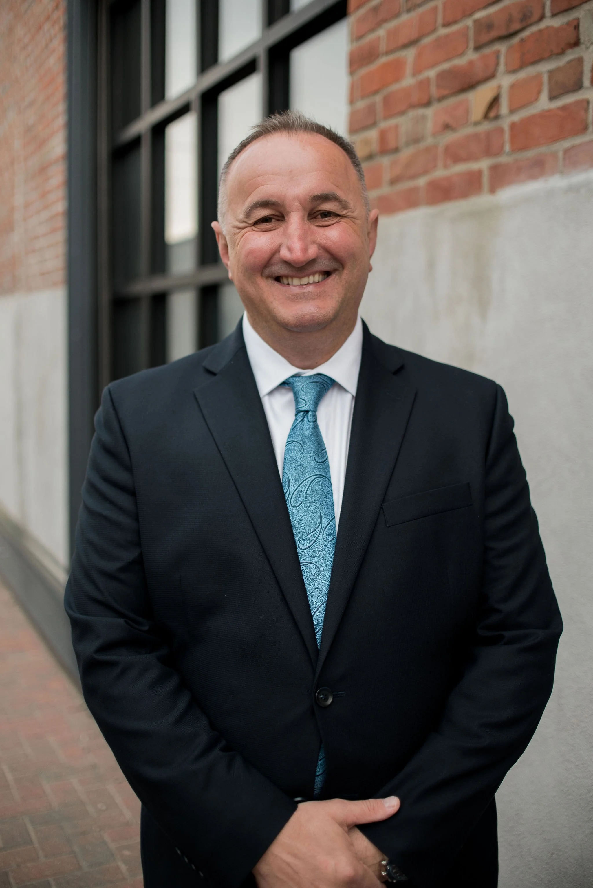 A man in a black suit, white shirt, and blue patterned tie smiling and standing outdoors against a brick wall and window.