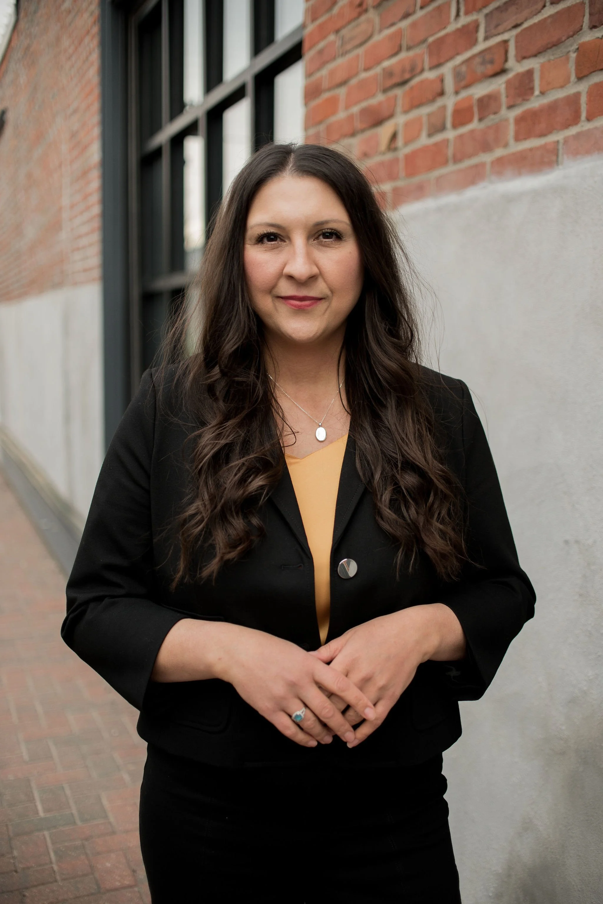A woman with long dark hair wearing a black blazer and yellow top, standing outdoors against a brick wall with black window frames, arms crossed, facing camera.