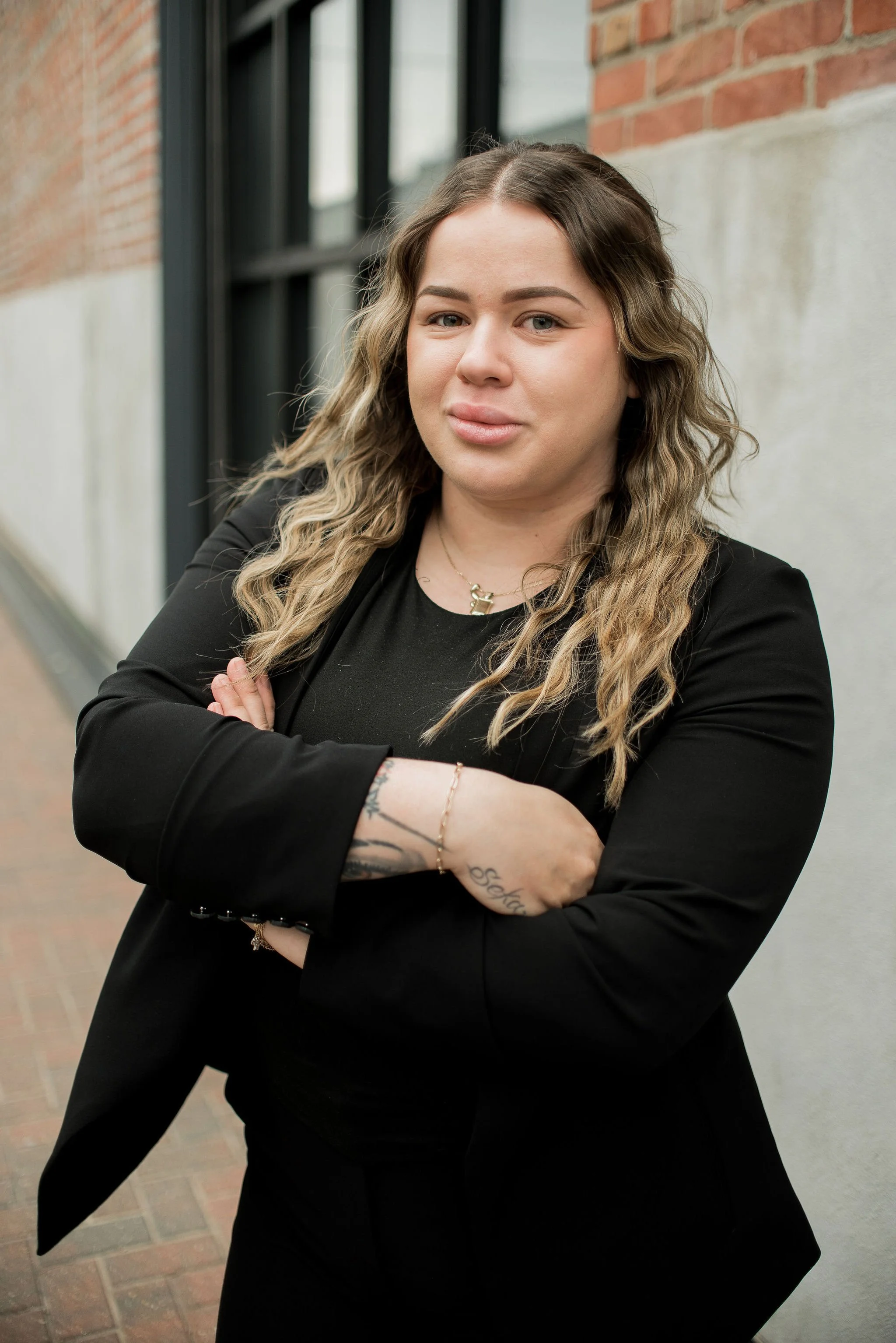 A young woman with long, wavy hair, wearing a black blazer and t-shirt, standing against an urban brick wall, with her arms crossed and a confident expression.