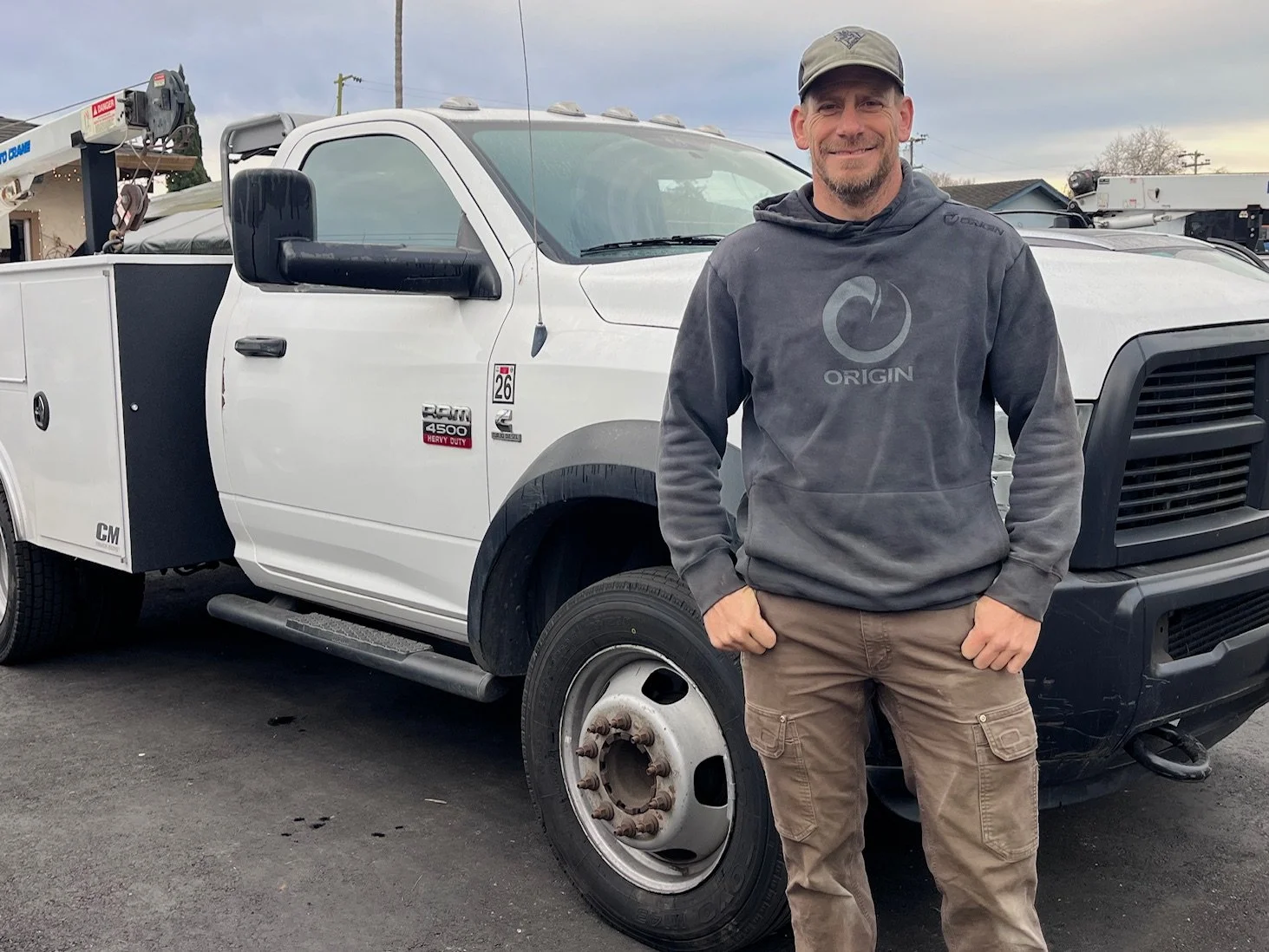 A man standing in front of a white utility truck, smiling, wearing a gray hoodie and khaki pants, with a construction crane visible in the background.
