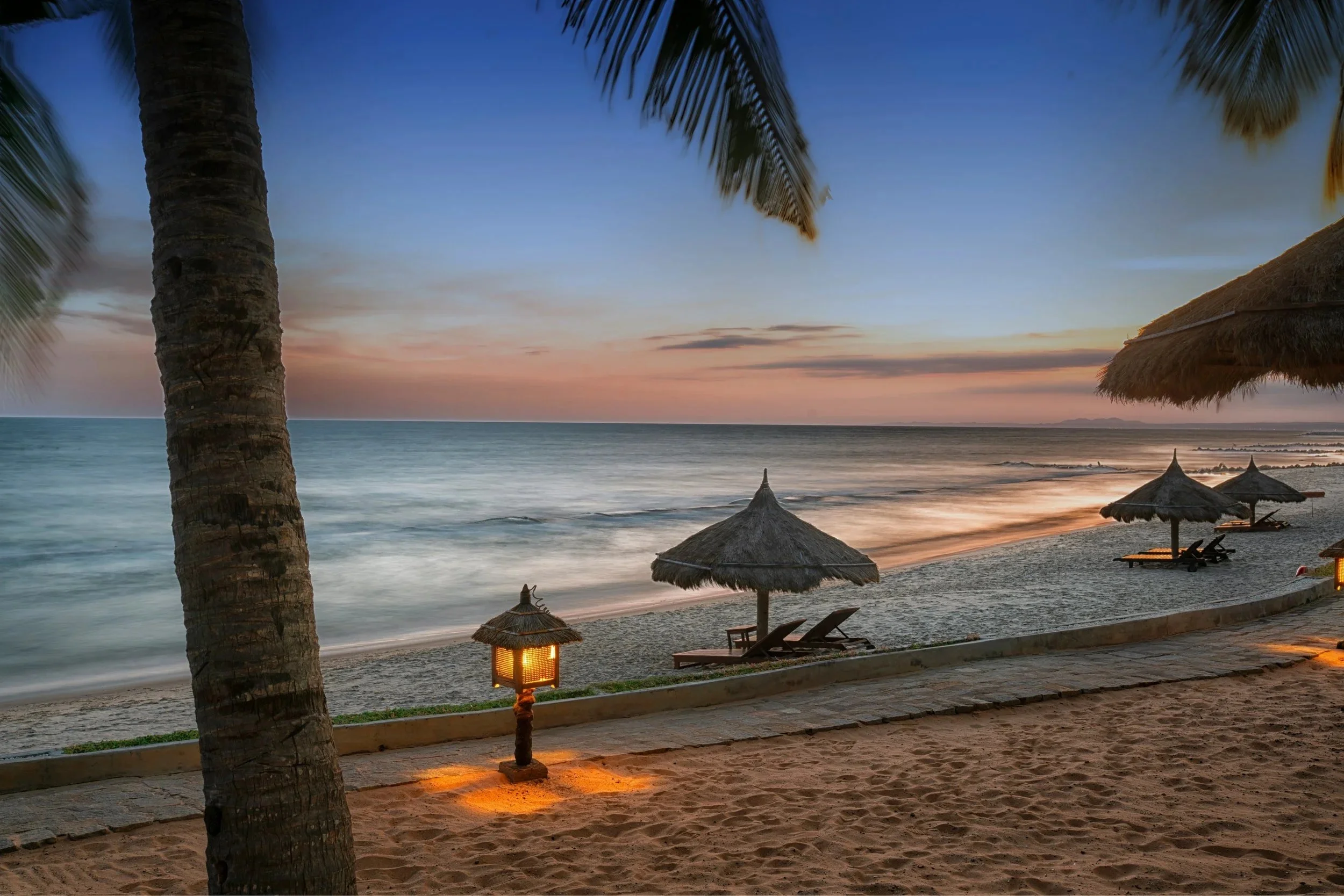 Sandy Beach with a small boardwalk lit my latern light in the foreground with a sandy beach, a palapa with beach chairs and ocean in the background, all under a clear sunset sky