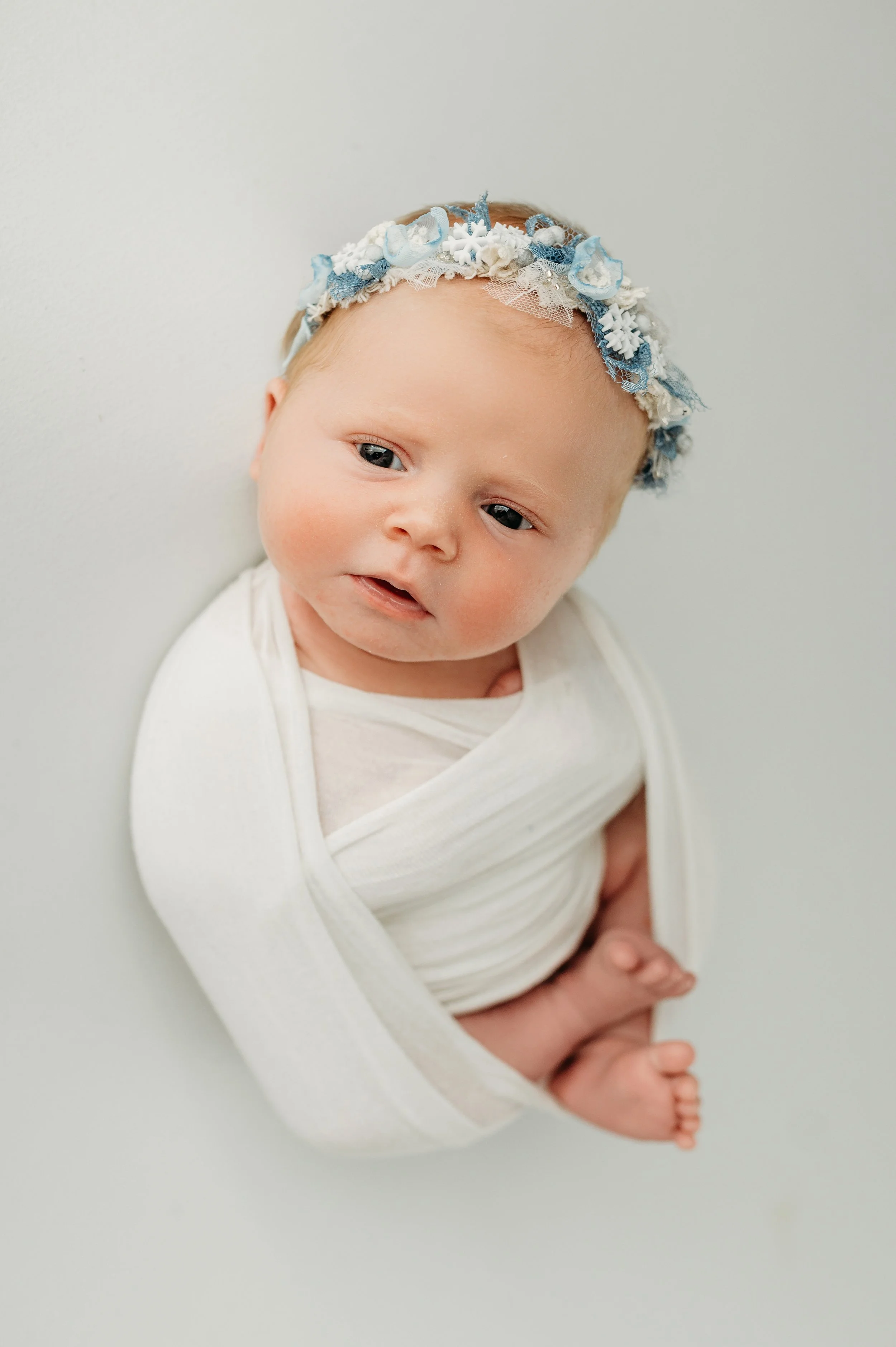 A baby with blue eyes and light skin, wearing a white outfit and a decorative headband with blue and white lace and fabric, is lying on its side on a plain light-colored surface.
