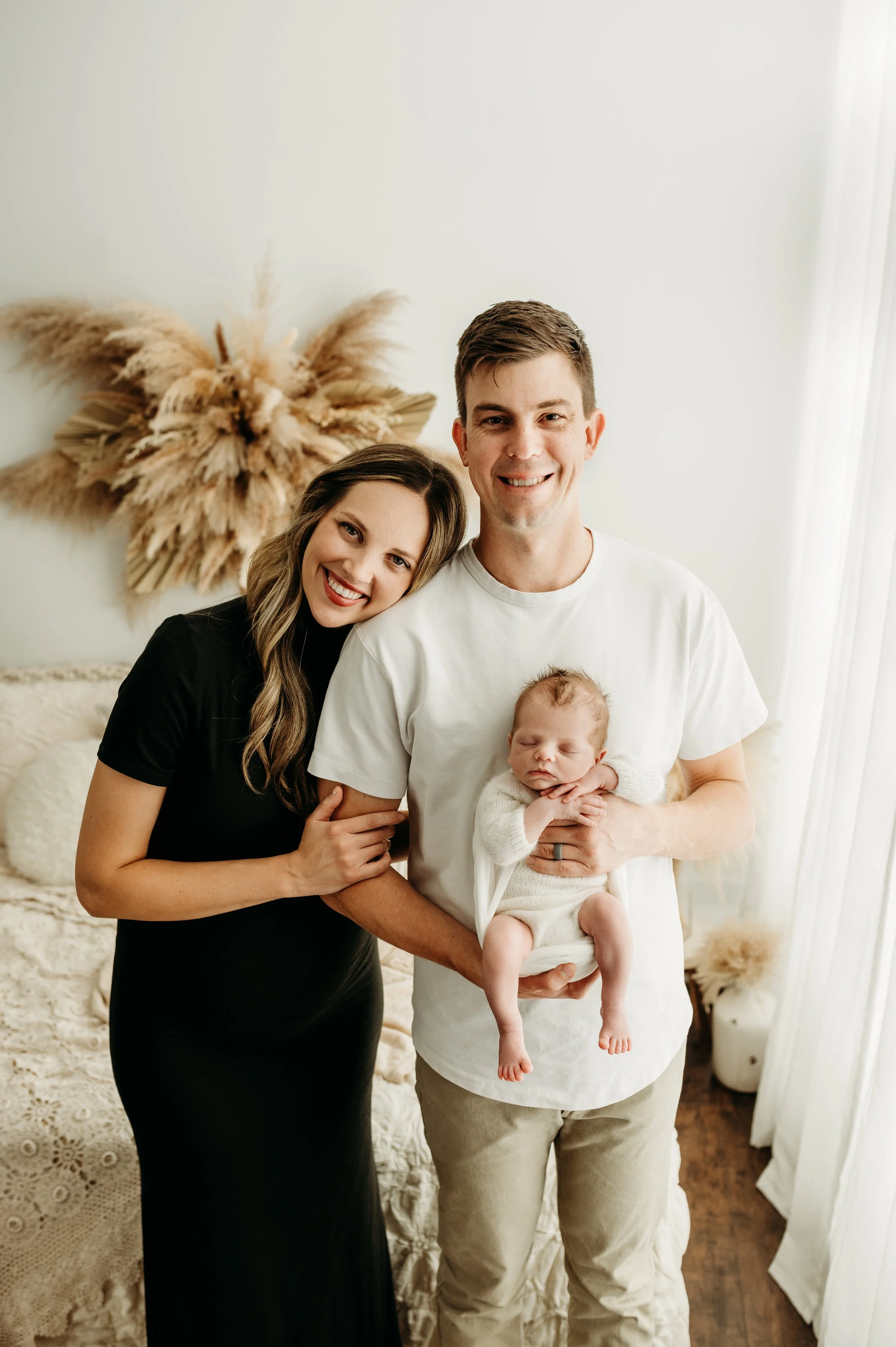 A smiling couple, a woman with long wavy brown hair wearing a black dress and a man with short brown hair in a white t-shirt and beige pants holding a sleeping baby in a cream-colored outfit, standing in a bright room with a dried flower wall art and a cozy bed with textured bedding.