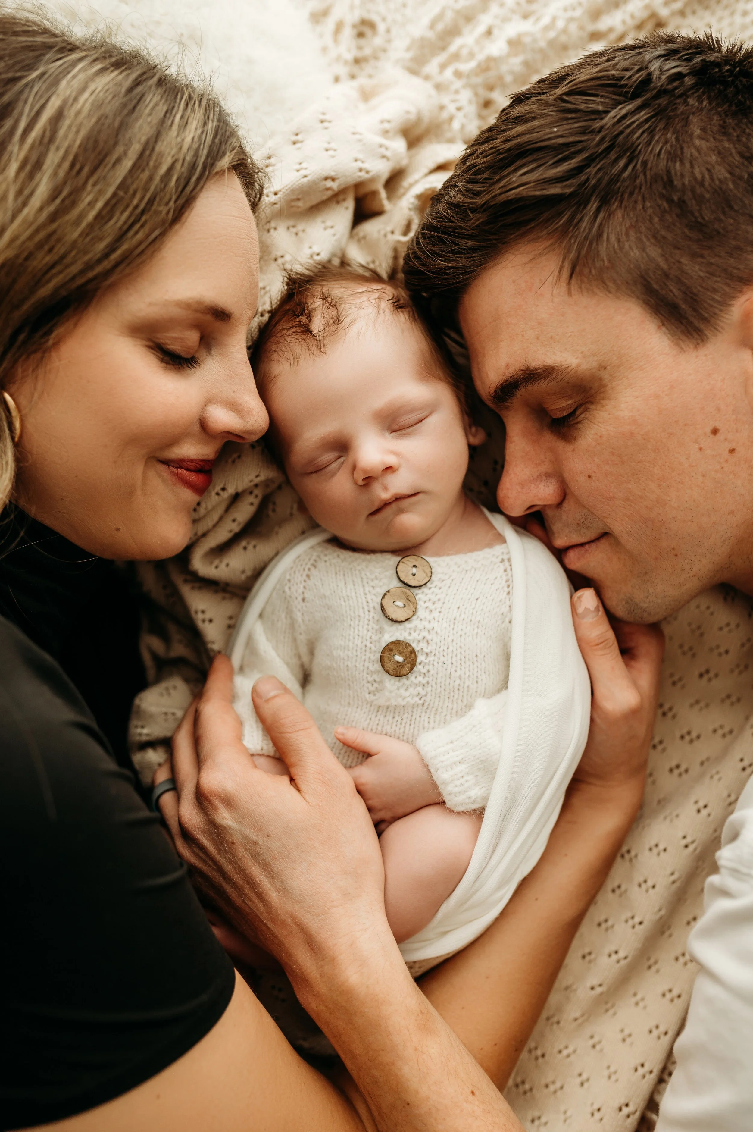 A family of three, including a woman, a man, and a baby, all with their eyes closed, lying together on a soft, cream-colored blanket, sharing an intimate moment of closeness and affection.