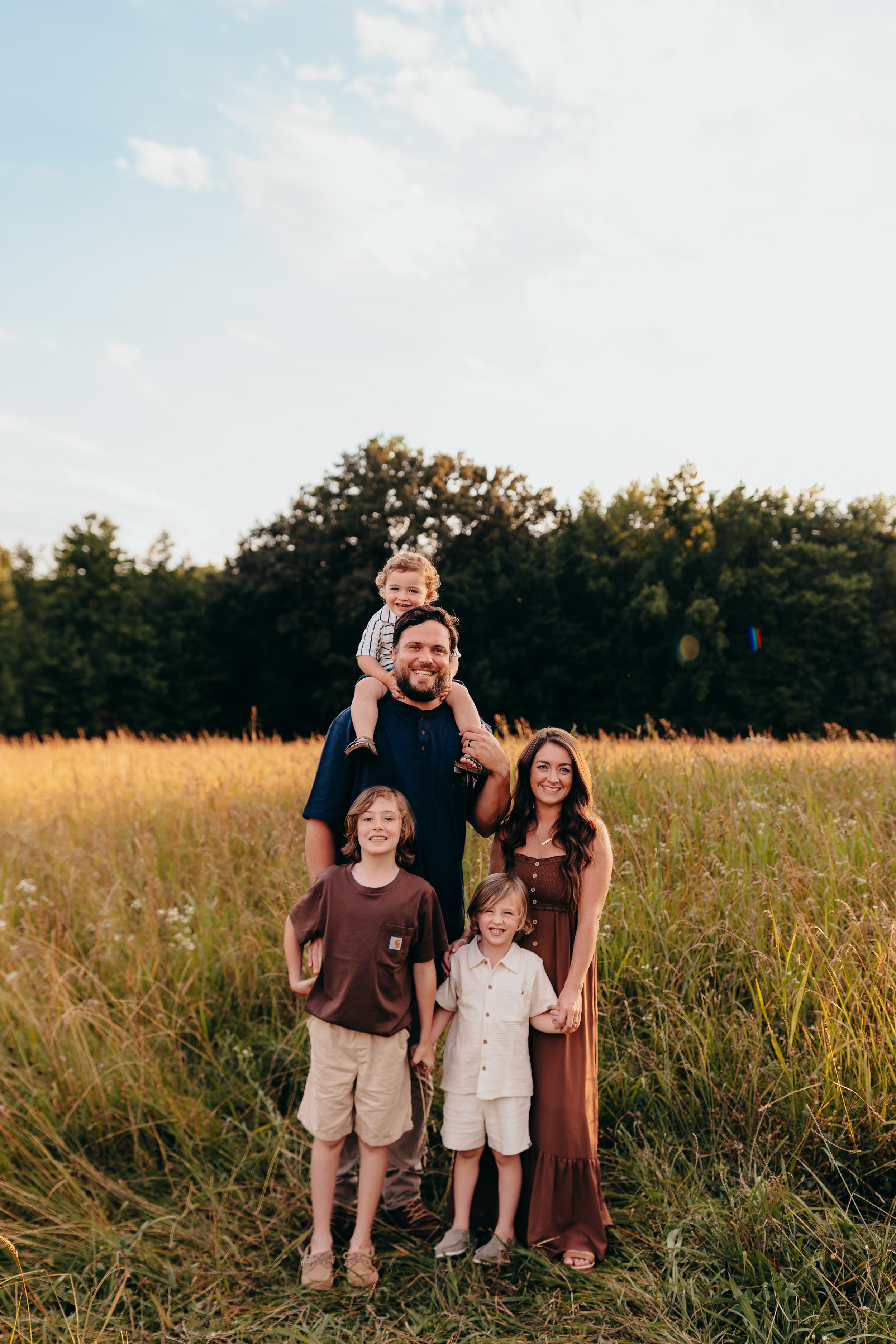 Family of five, including father, mother, and three children, standing in tall grass meadow with trees and blue sky in the background.