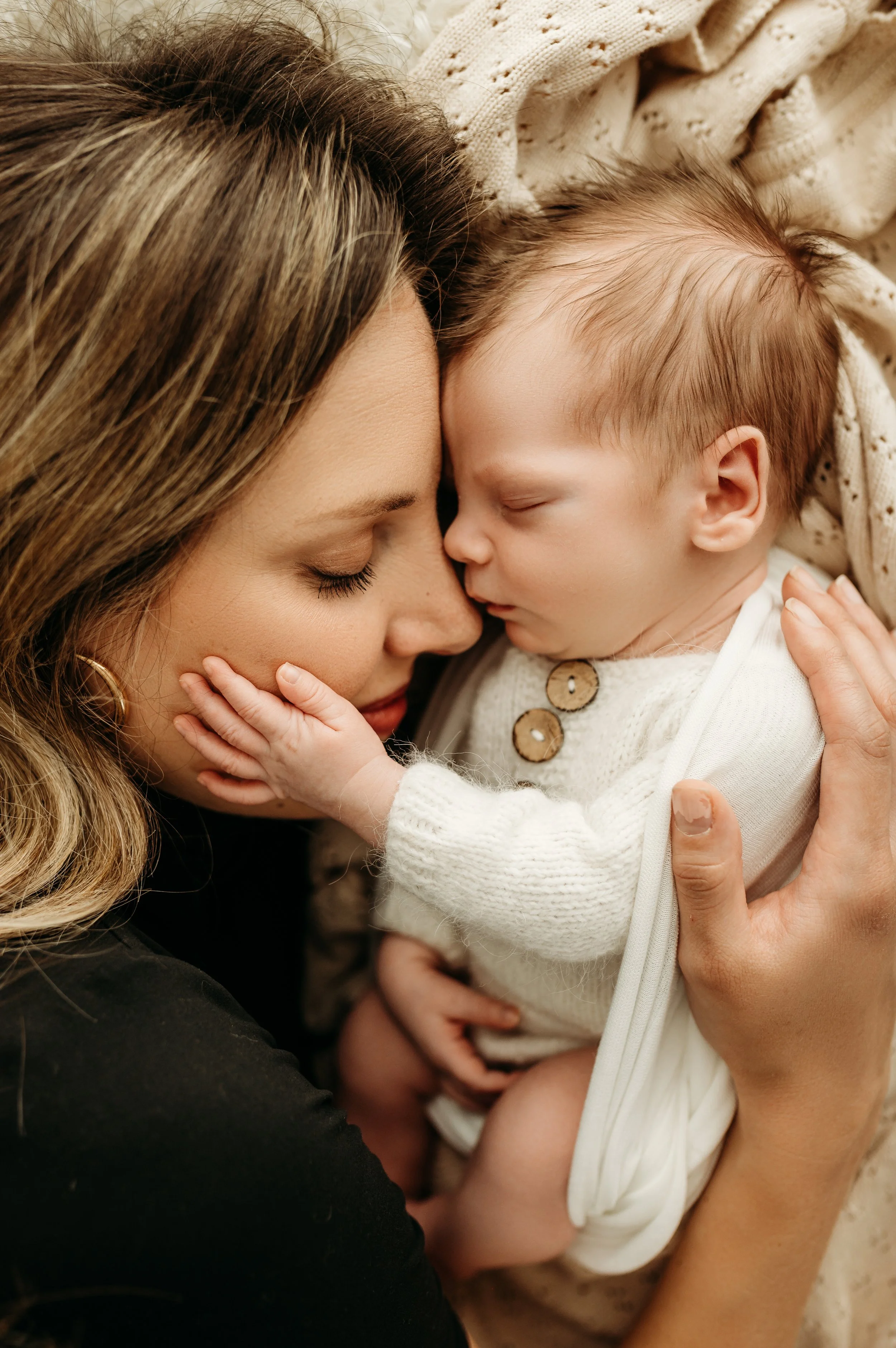 A woman with long light brown hair and closed eyes gently cradles a sleeping baby with closed eyes and light brown hair. They are close together, pressed foreheads, sharing an intimate moment, with a warm beige knitted blanket in the background.
