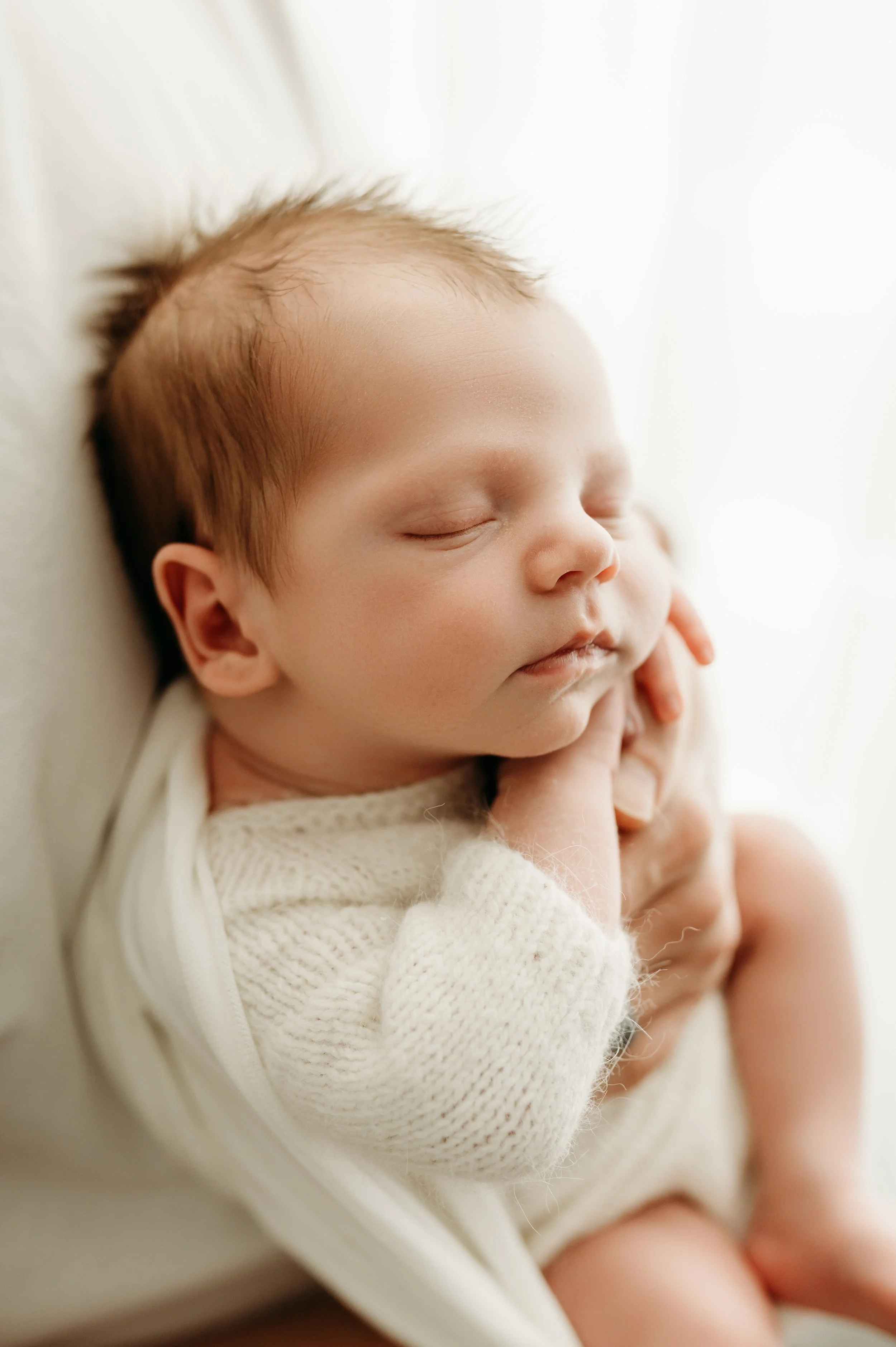 A sleeping baby with fair skin, closed eyes, and light brown hair, resting against an adult's arm and hand, wearing a cozy white knit sweater.