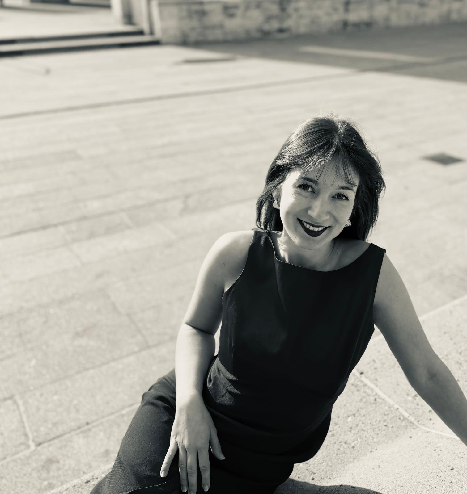 Schwarz-weiße Fotografie einer lächelnden Frau in schwarzen Kleid, sitzend auf einer Stufe, im Freien, mit einem offenen, freundlichen Ausdruck.