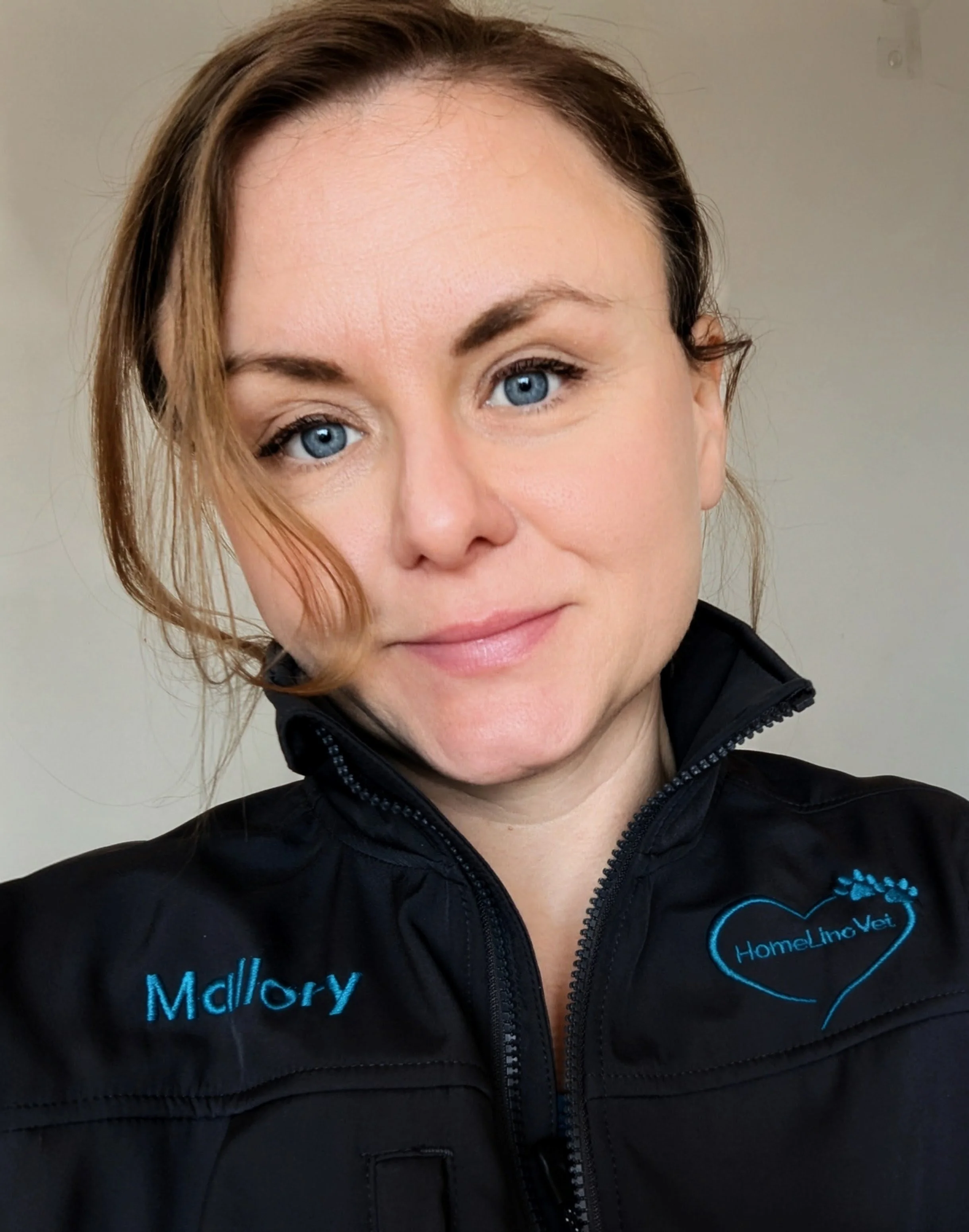 Close-up of a woman's face with fair skin, blue eyes, and light brown hair, wearing a black jacket with a heart-shaped logo and the name 'McIlroy' embroidered on it.