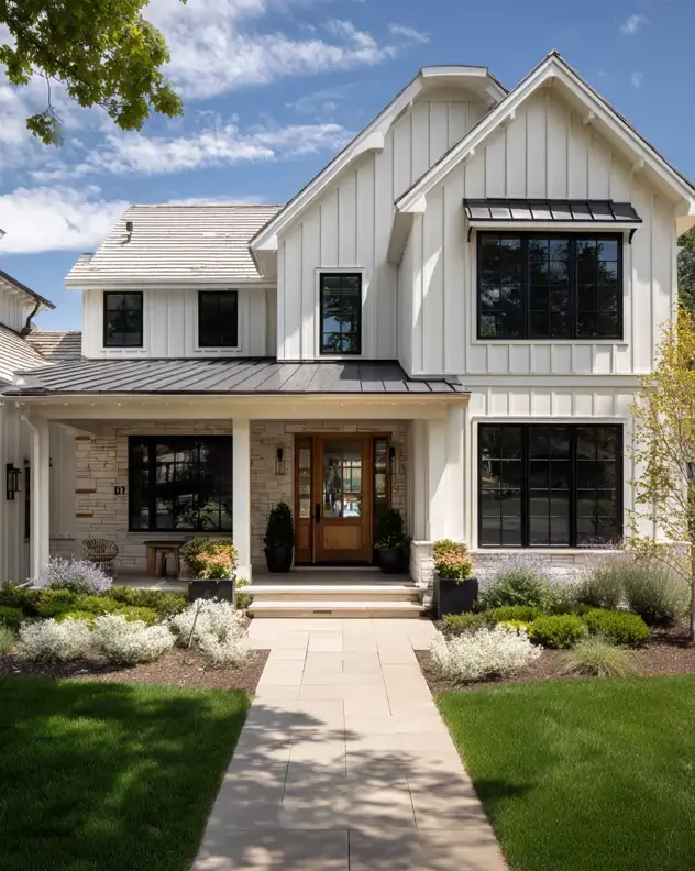 Modern two-story white house with black window frames and a wooden front door, surrounded by a landscaped yard with green grass and bushes, under a partly cloudy sky.