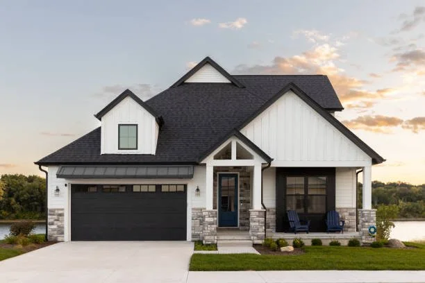 Modern two-story house with white siding, black roof, and attached two-car garage, featuring a small front porch with two chairs and a well-maintained lawn.