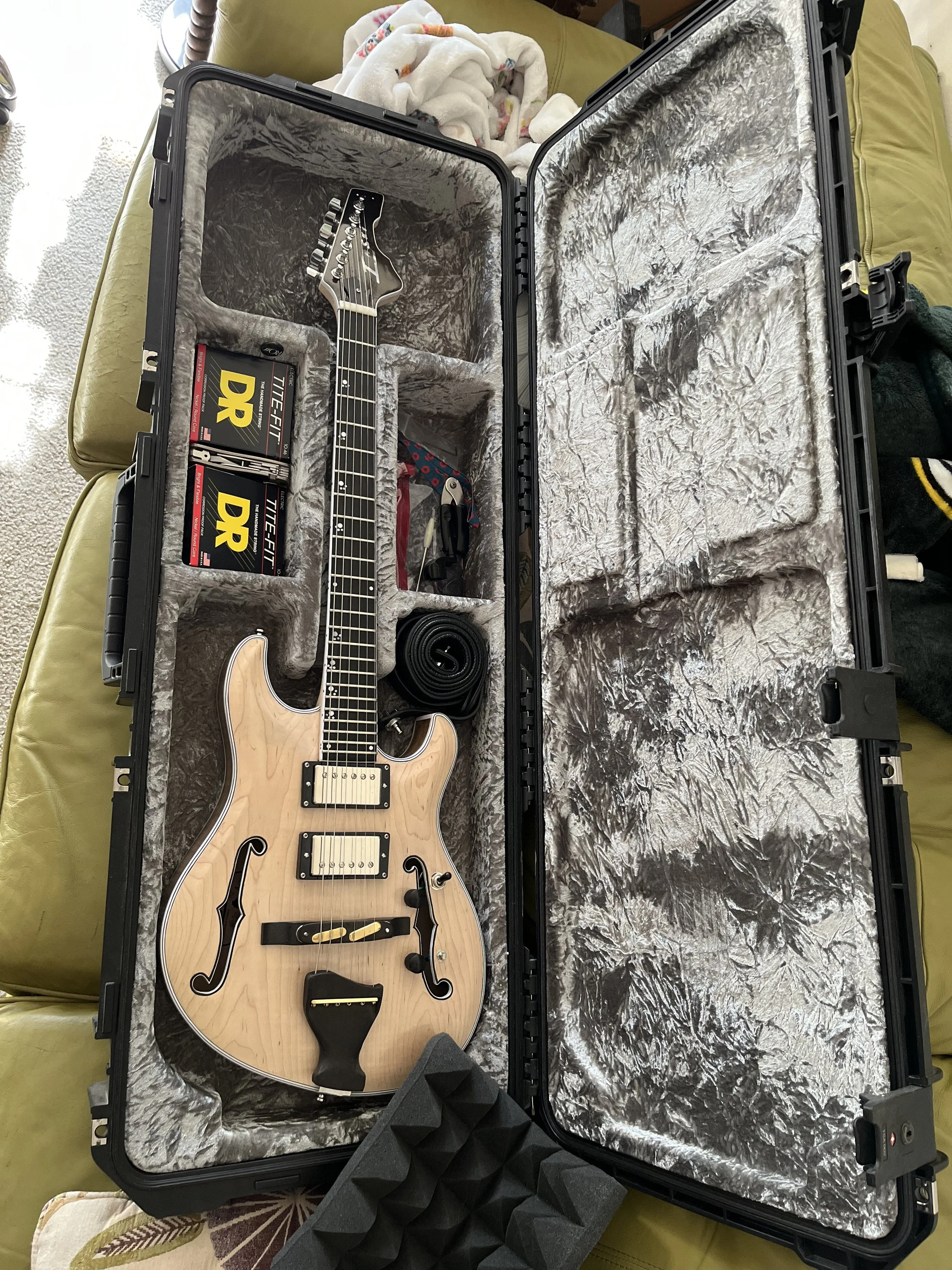 A semi-hollow electric guitar with a natural wood finish inside a hard protective case. The case has gray plush lining and is placed on a yellow sofa. There are two packs of DR Tune-Fit strings and some other accessories inside the case.