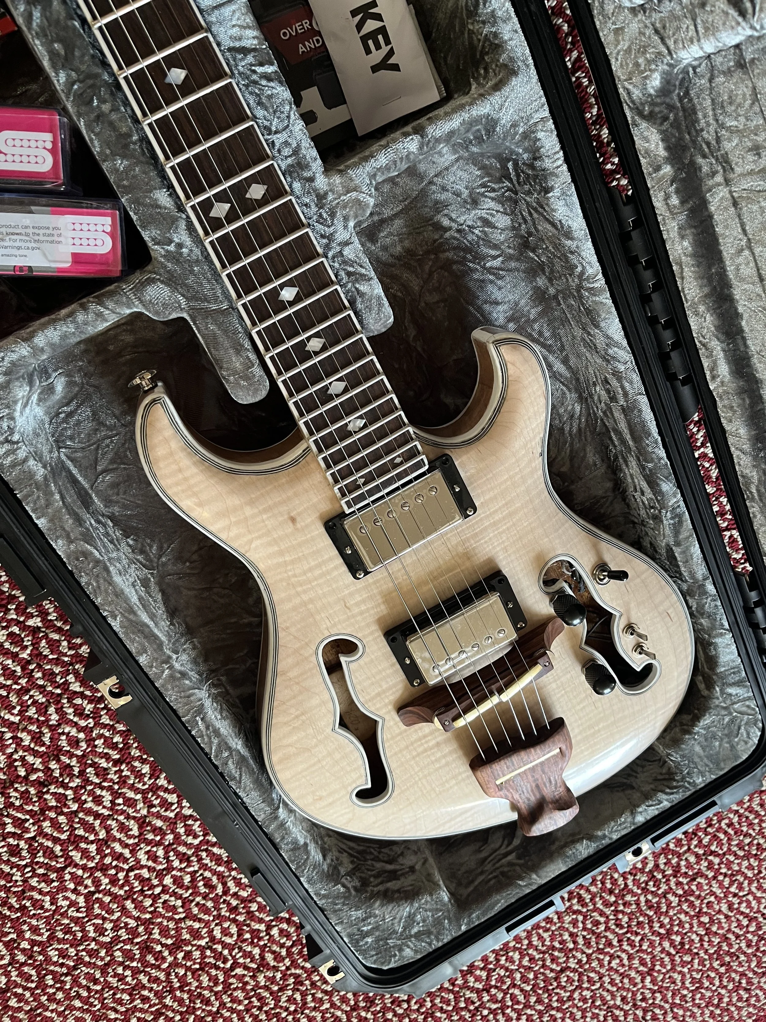 An electric guitar with a natural wood finish resting in a black hard case lined with silver interior material. The guitar has a unique built-in sound hole, metallic pickups, and a wooden control knob. Various boxes and supplies are visible around th