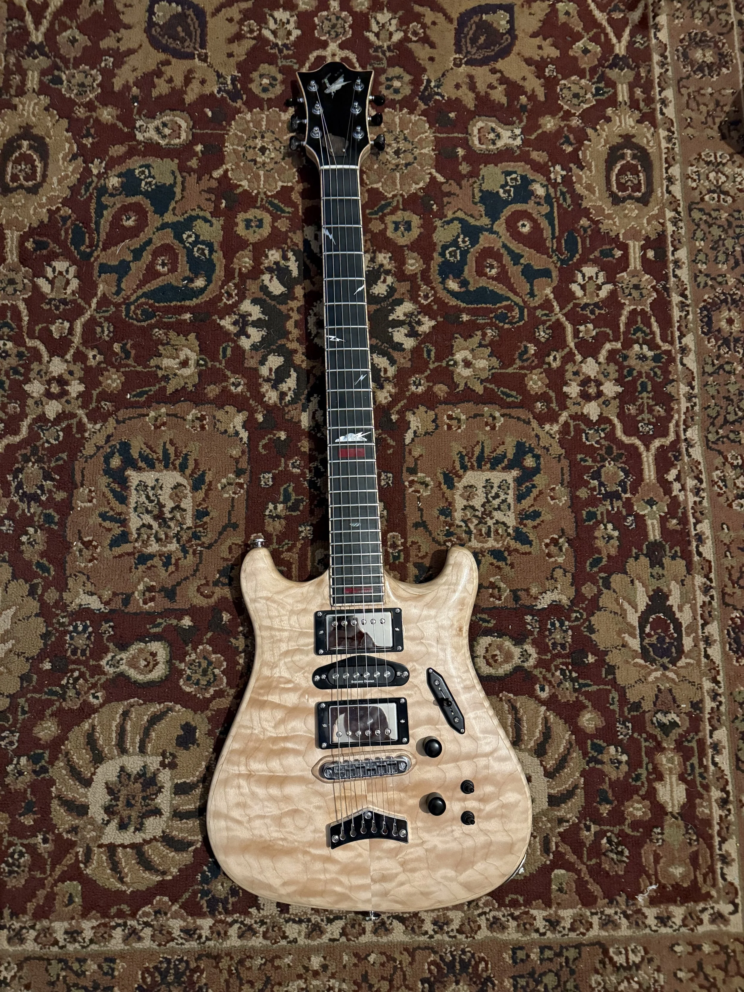 A light-colored electric guitar with a quilted maple top, black hardware, and a black pickguard, lying on a patterned red, beige, and blue rug.