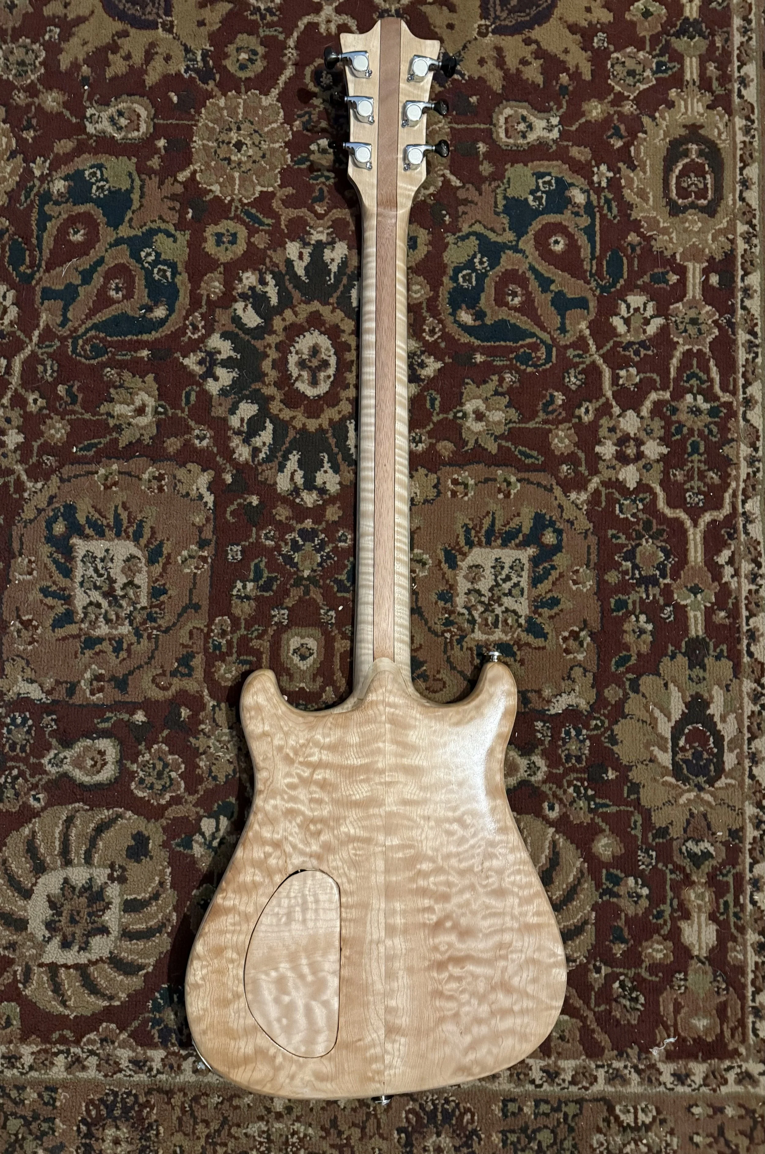 Back of a unfinished electric guitar body and neck with tuning pegs, placed on a patterned area rug.