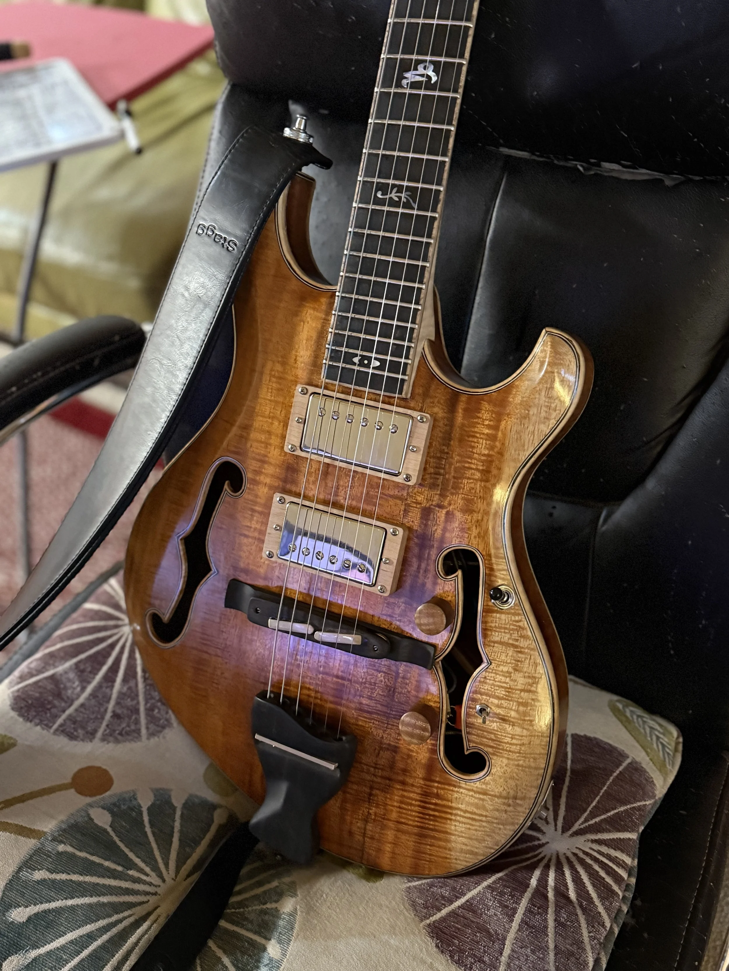 An electric guitar with a wooden finish resting on a cushion with a floral pattern. The guitar has a semi-hollow body with f-holes, a black strap, and chrome hardware.