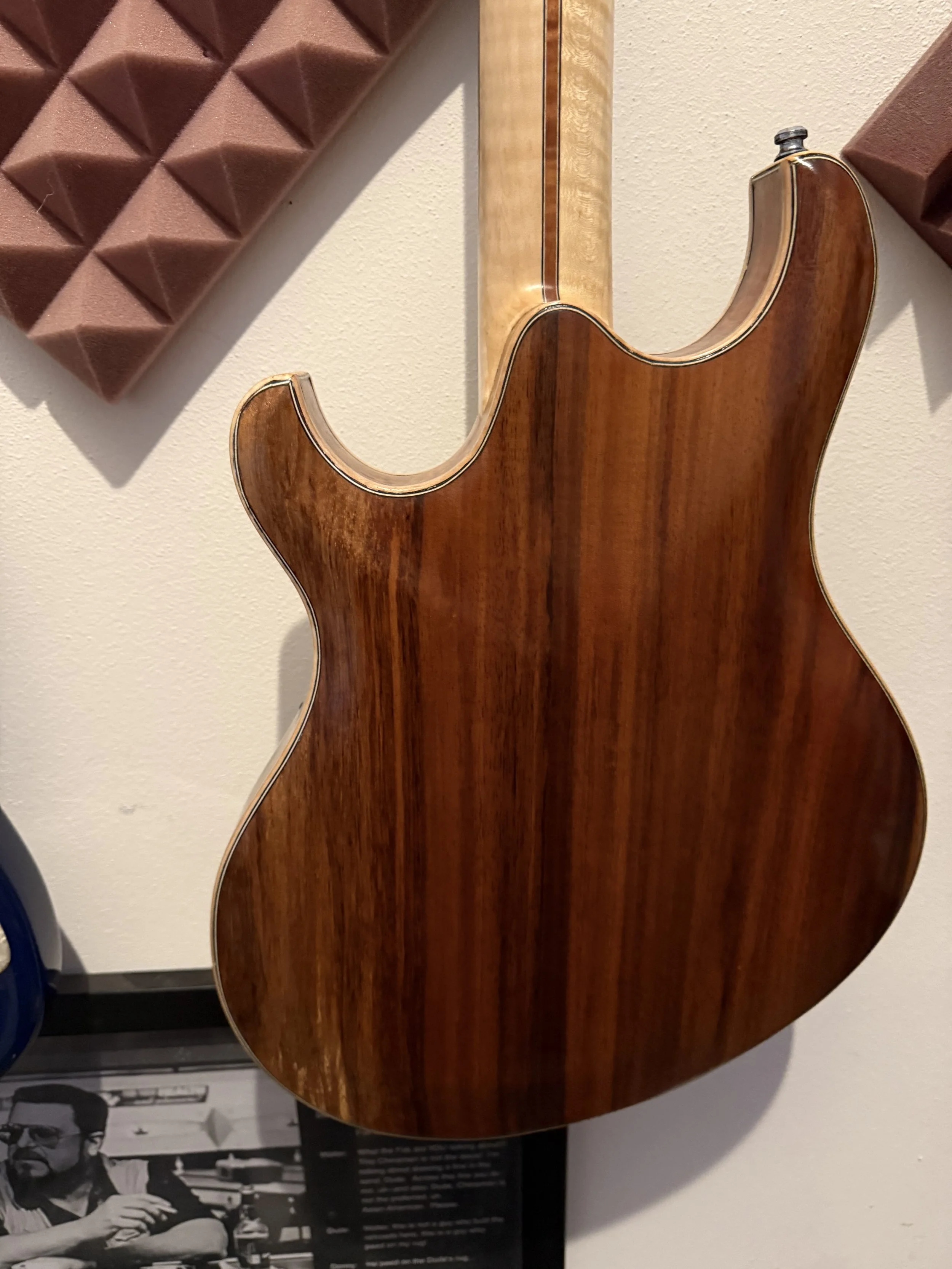 Close-up of the back of an electric guitar with wood grain finish, mounted on a beige wall. Part of a framed black and white photo is visible at the bottom.