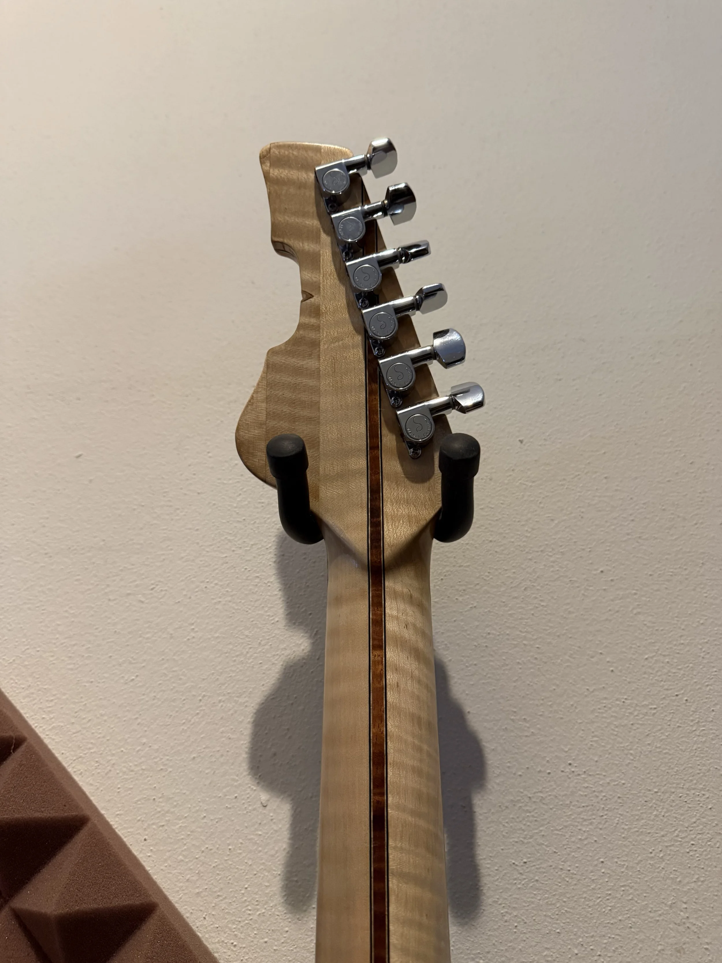 Back of a guitar headstock with tuning pegs against a beige wall, with brown staircase in the lower corner.