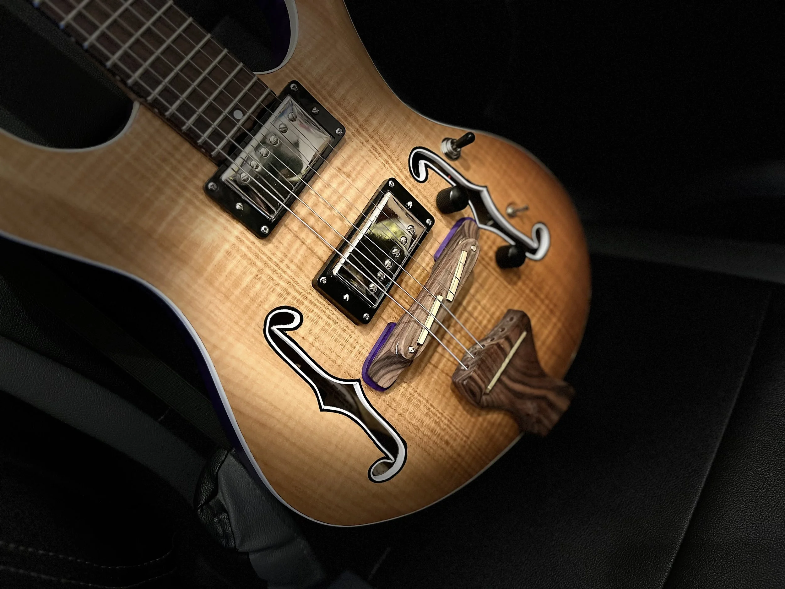 Close-up of a light-colored wooden electric guitar with black and white stickers, resting on a black surface.