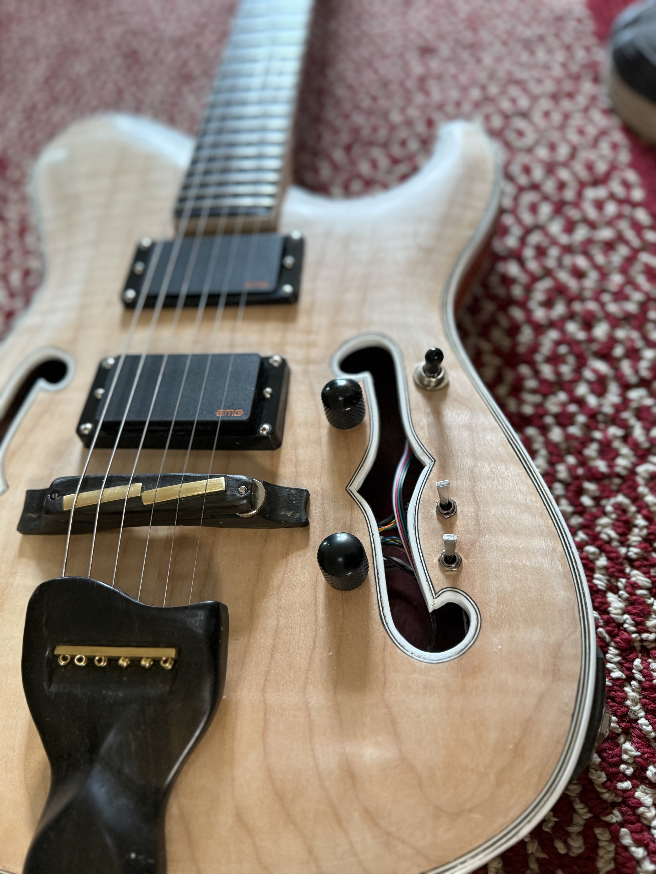 A close-up of an electric guitar with a light-colored wooden body, black pickups, control knobs, and a black pickguard, resting on a red patterned fabric.