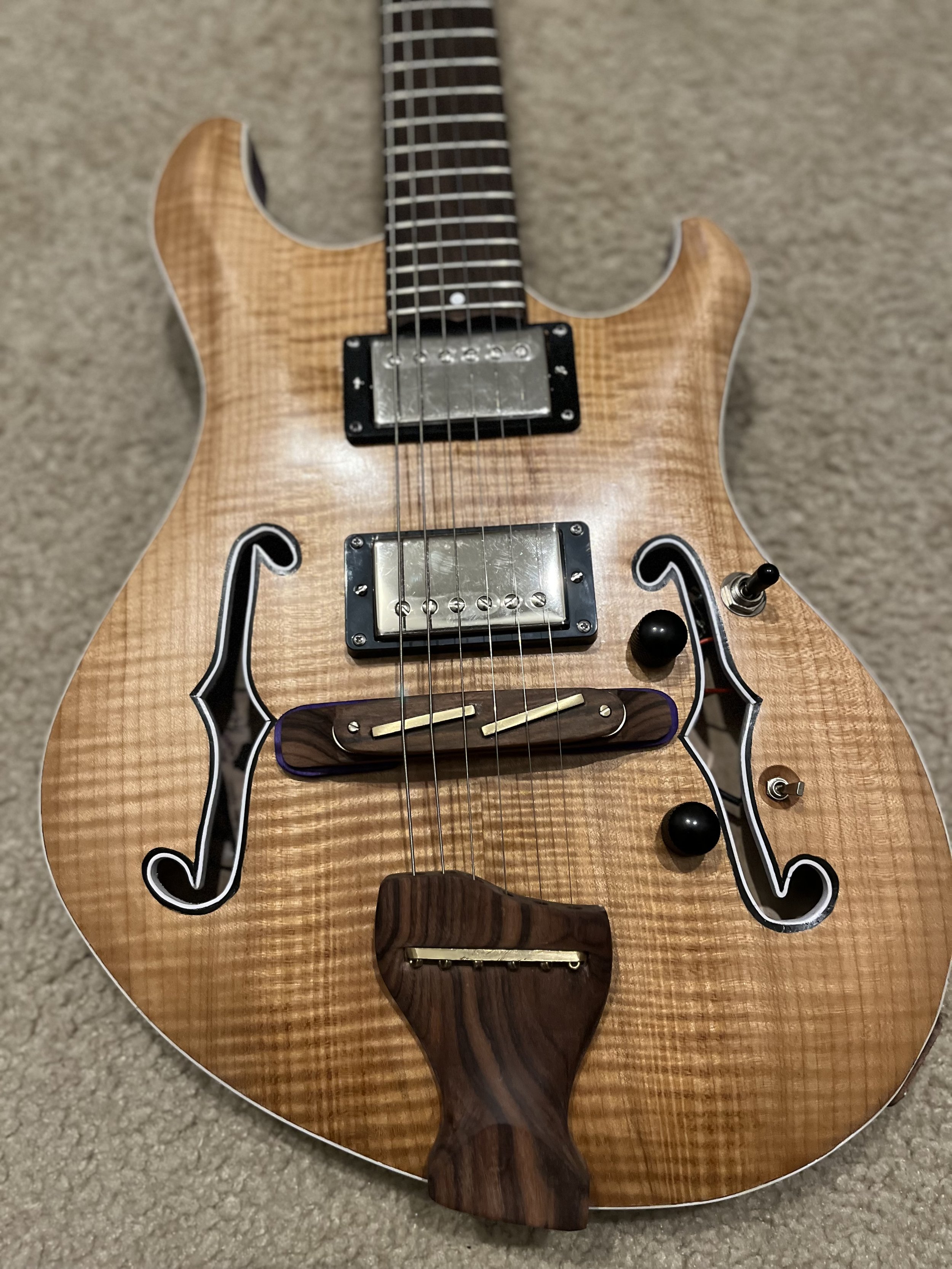 Close-up of a semi-acoustic electric guitar with a natural wood finish, decorative black and white binding, and a wooden bridge, lying on a beige carpet.