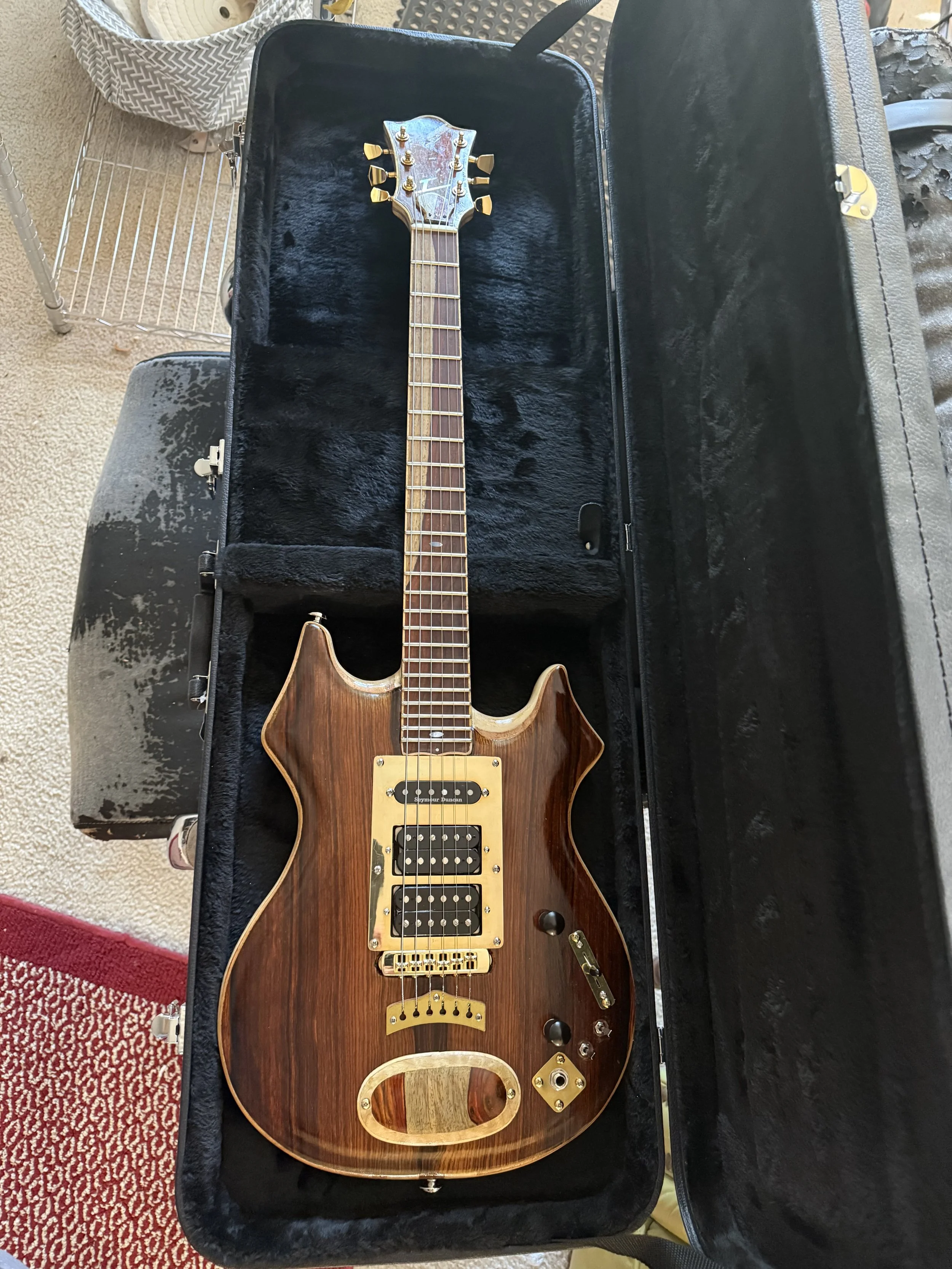 An electric guitar with a wooden body, gold hardware, and multiple pickups, resting in an open black case lined with plush fabric.