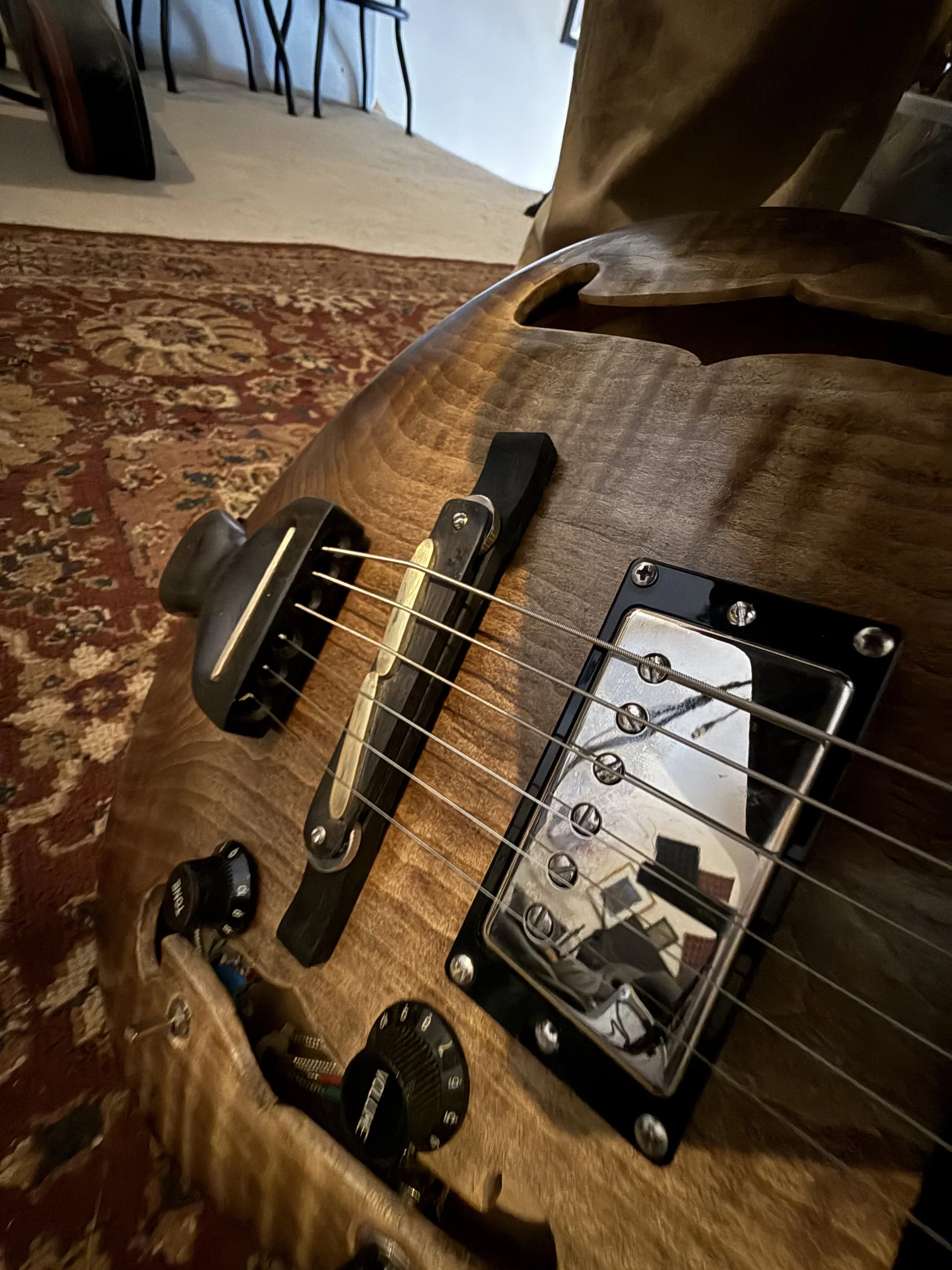 A close-up of a dark wooden electric guitar laying on a patterned area rug, with parts of a heel, bridge, strings, and pickups visible.