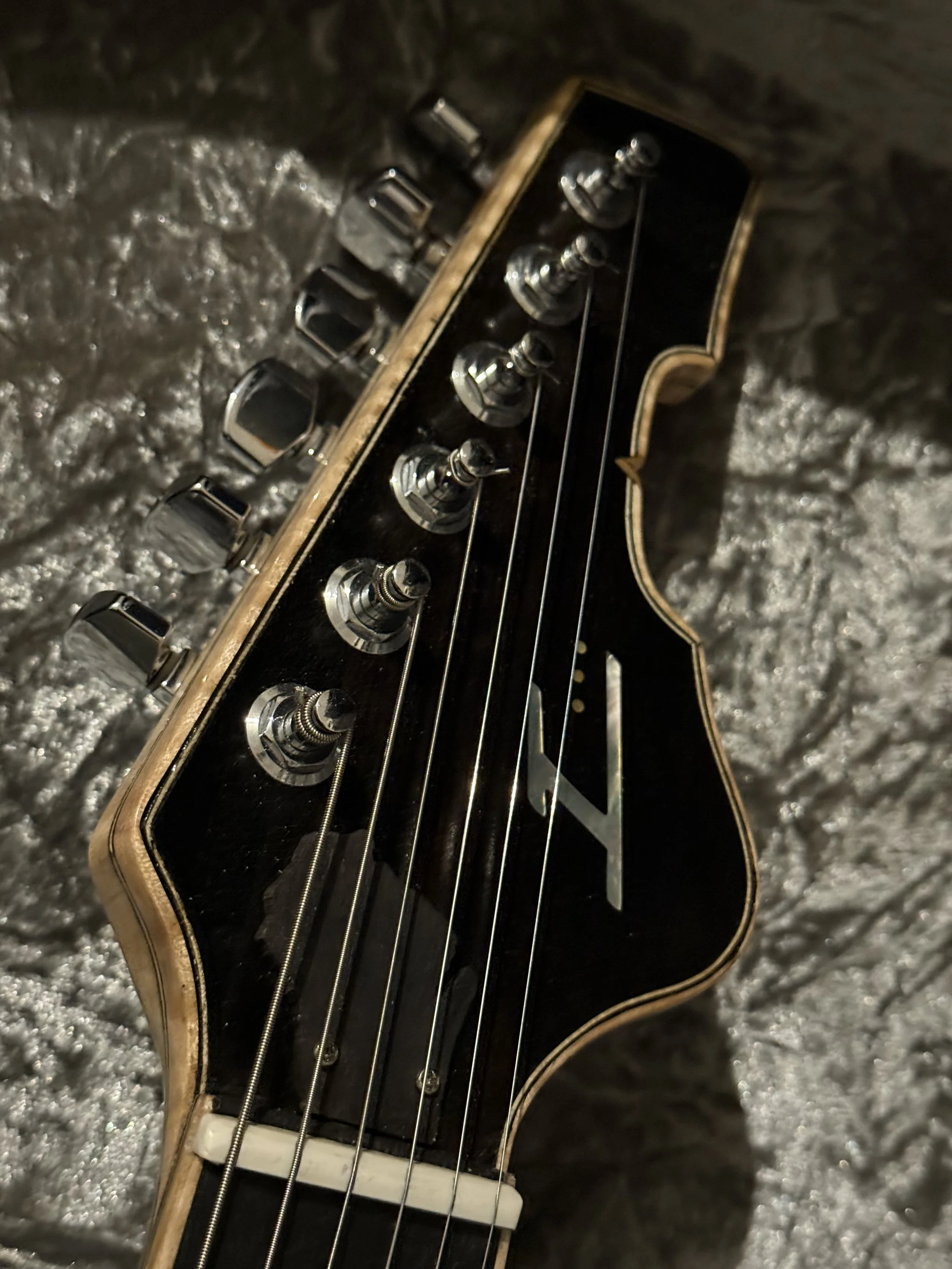 Close-up of an electric guitar neck with tuning pegs, strings, and a black pickguard, placed on a cushioned surface.