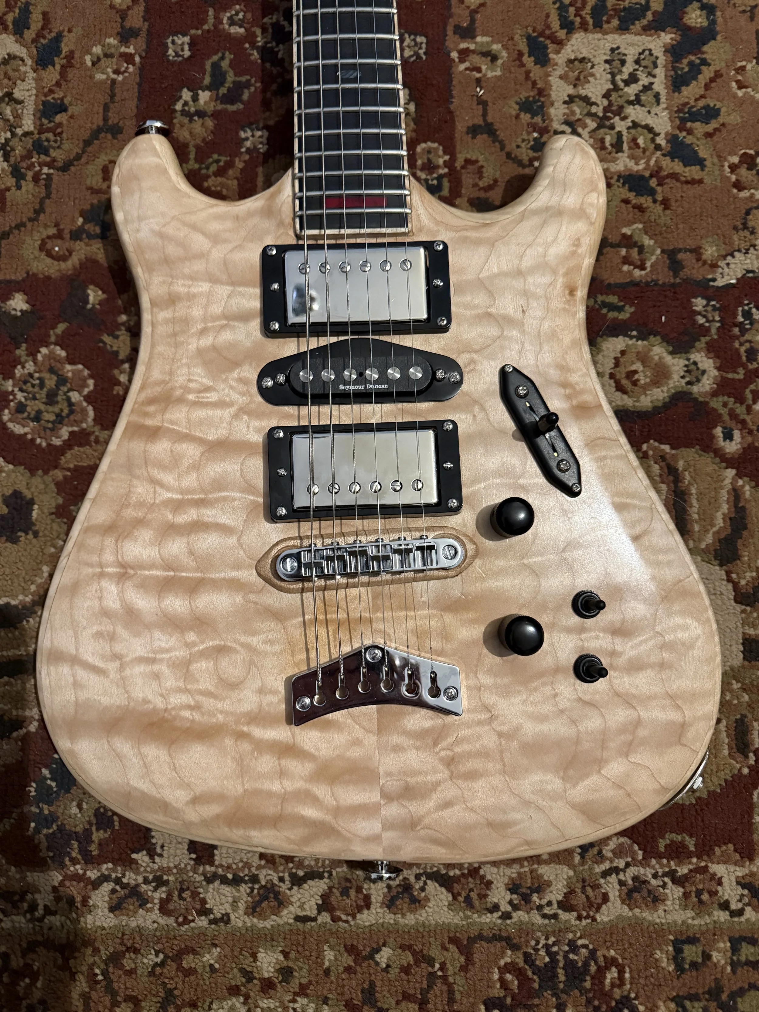 An electric guitar with a natural wood finish and quilted maple top, lying on a patterned rug.