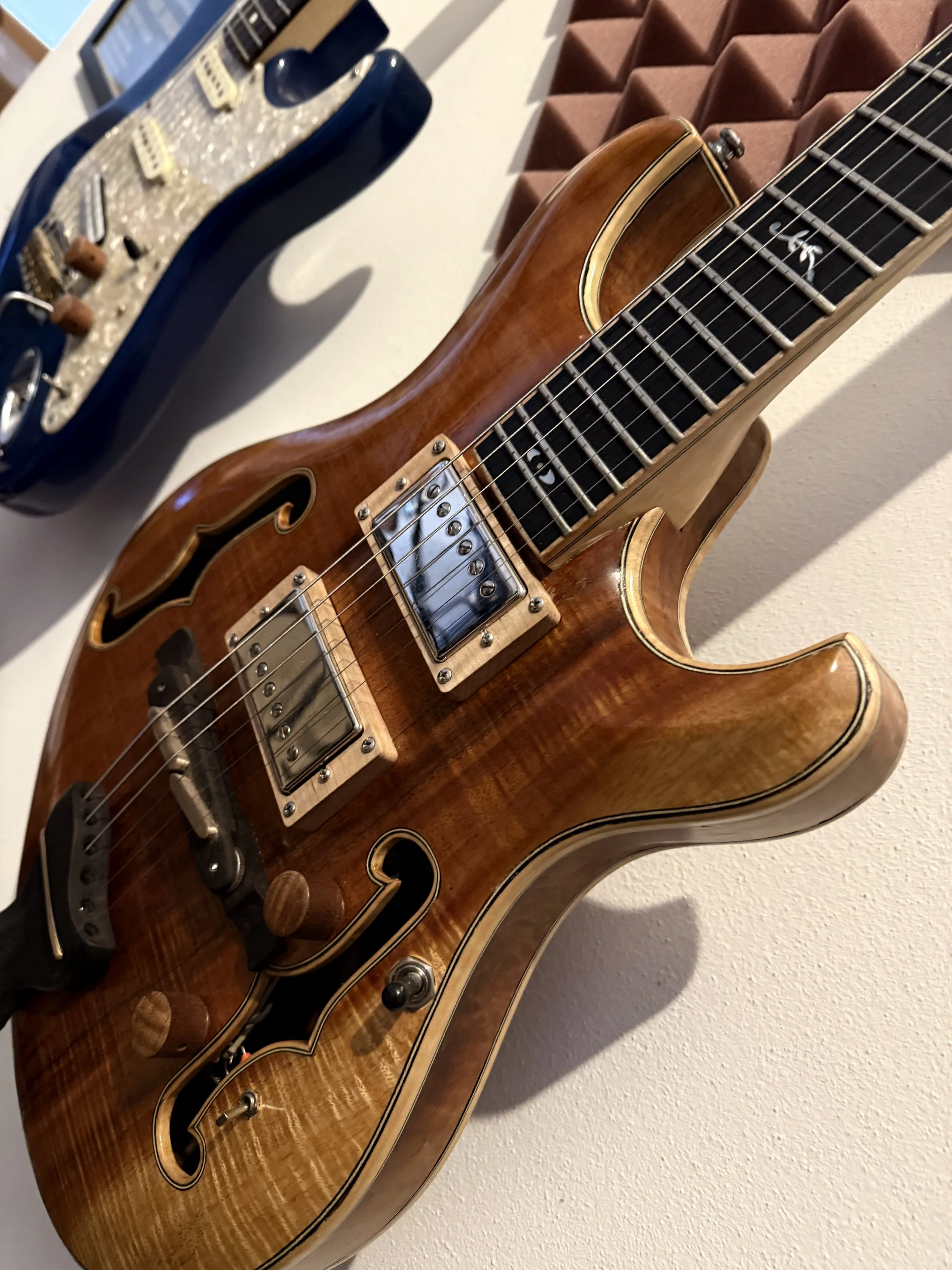 A wooden electric guitar with a hollow body and a black fretboard, placed against a beige wall. Next to it, there is a blue electric guitar with a pearl-like pickguard and control knobs.
