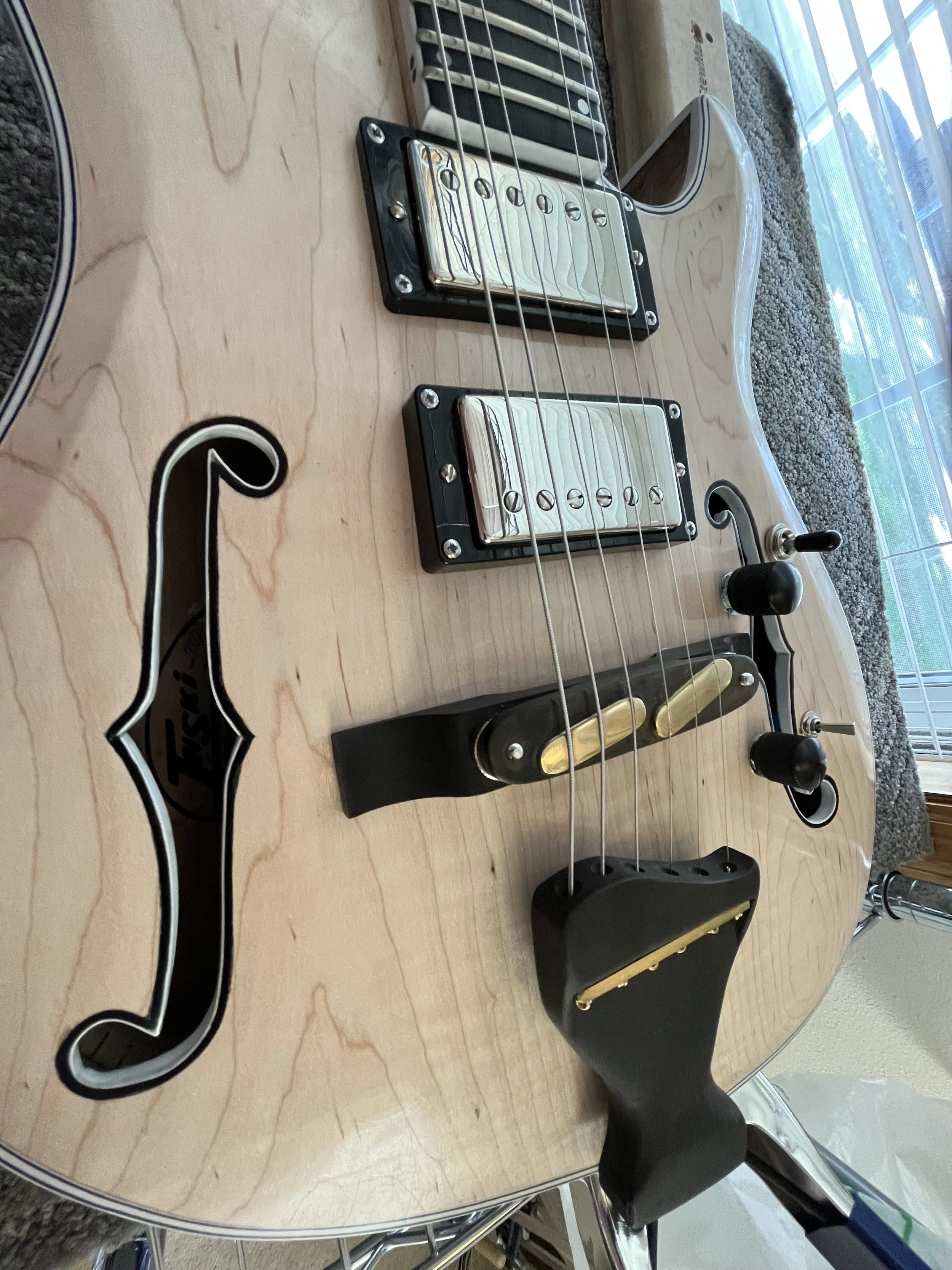 Close-up view of an electric guitar with a light wood finish, displaying strings, pickups, and parts of the bridge, with a black strap attached to the bottom.