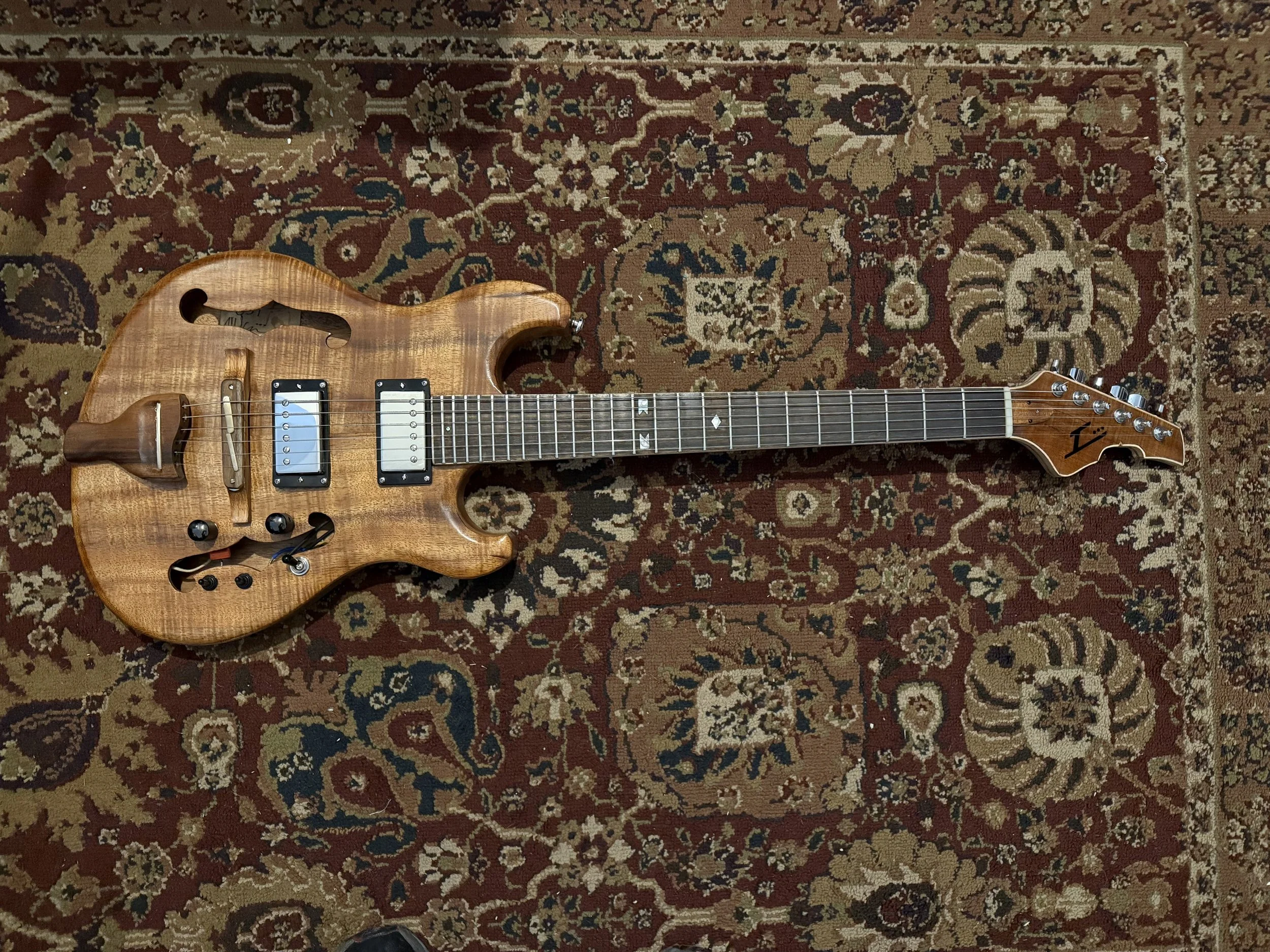 An electric guitar with a natural wood finish, black hardware, and a black bolt-on neck, resting on a patterned oriental rug.
