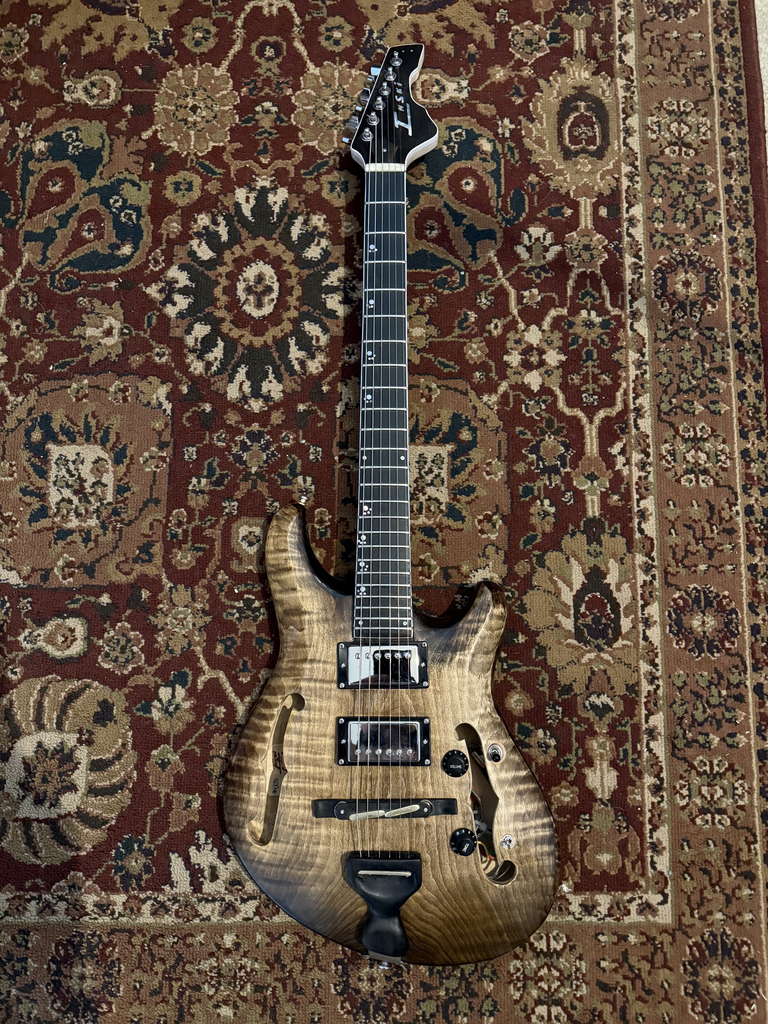 A semi-hollow electric guitar with a natural wood finish, black hardware, and F-hole design, lying on a patterned red and beige rug.