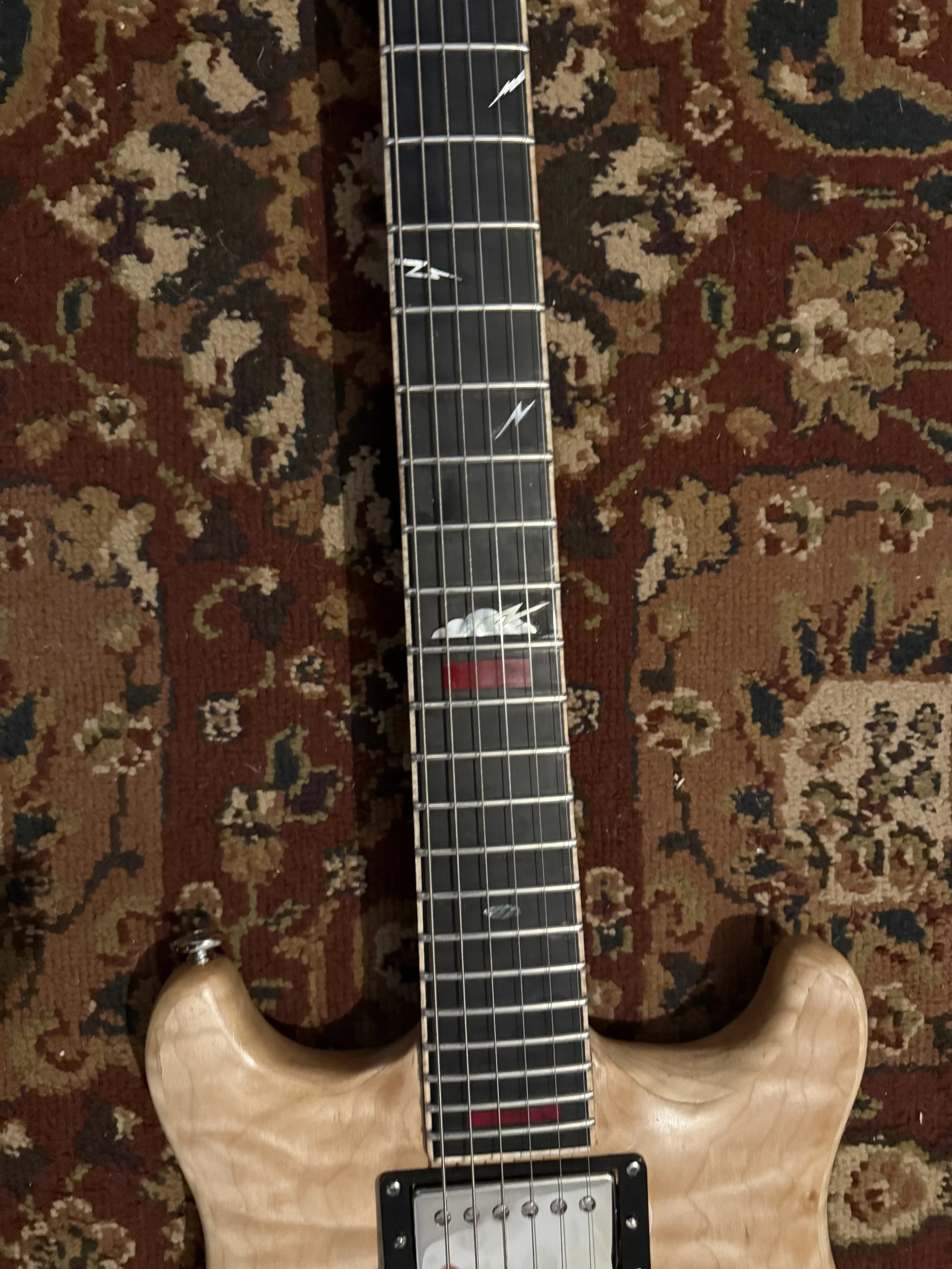 A wooden electric guitar neck with six strings, placed on a patterned carpet. The guitar has a natural wood color and a white inlay at the fret marker. The photo is taken from above, showing part of the guitar and the person's feet at the bottom.