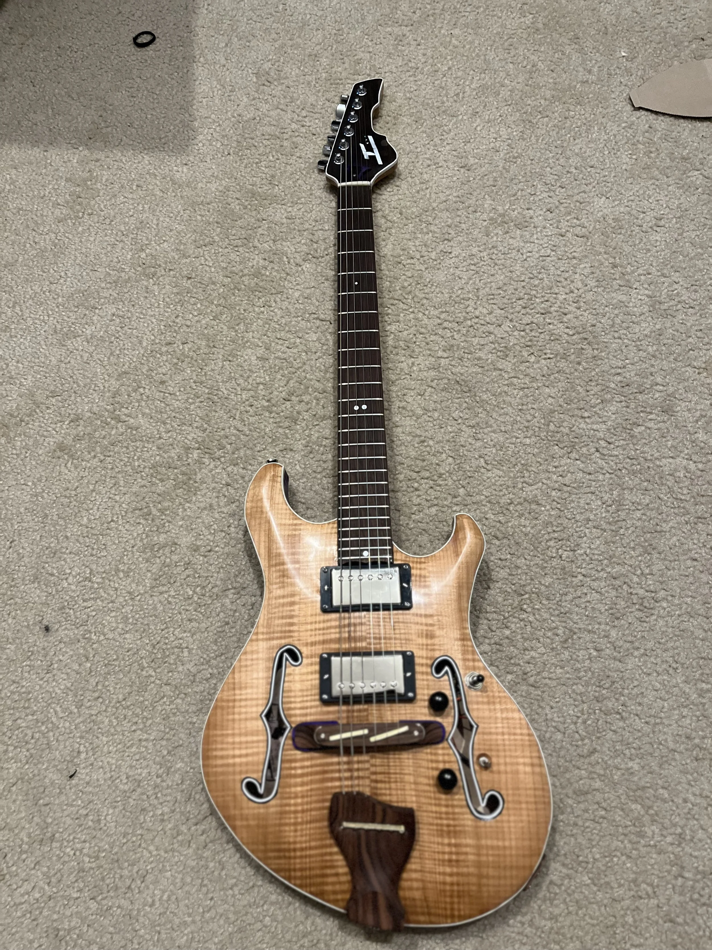 A semi-hollow electric guitar with a natural wood finish, black hardware, and decorative black and white binding on the body, lying on beige carpet.