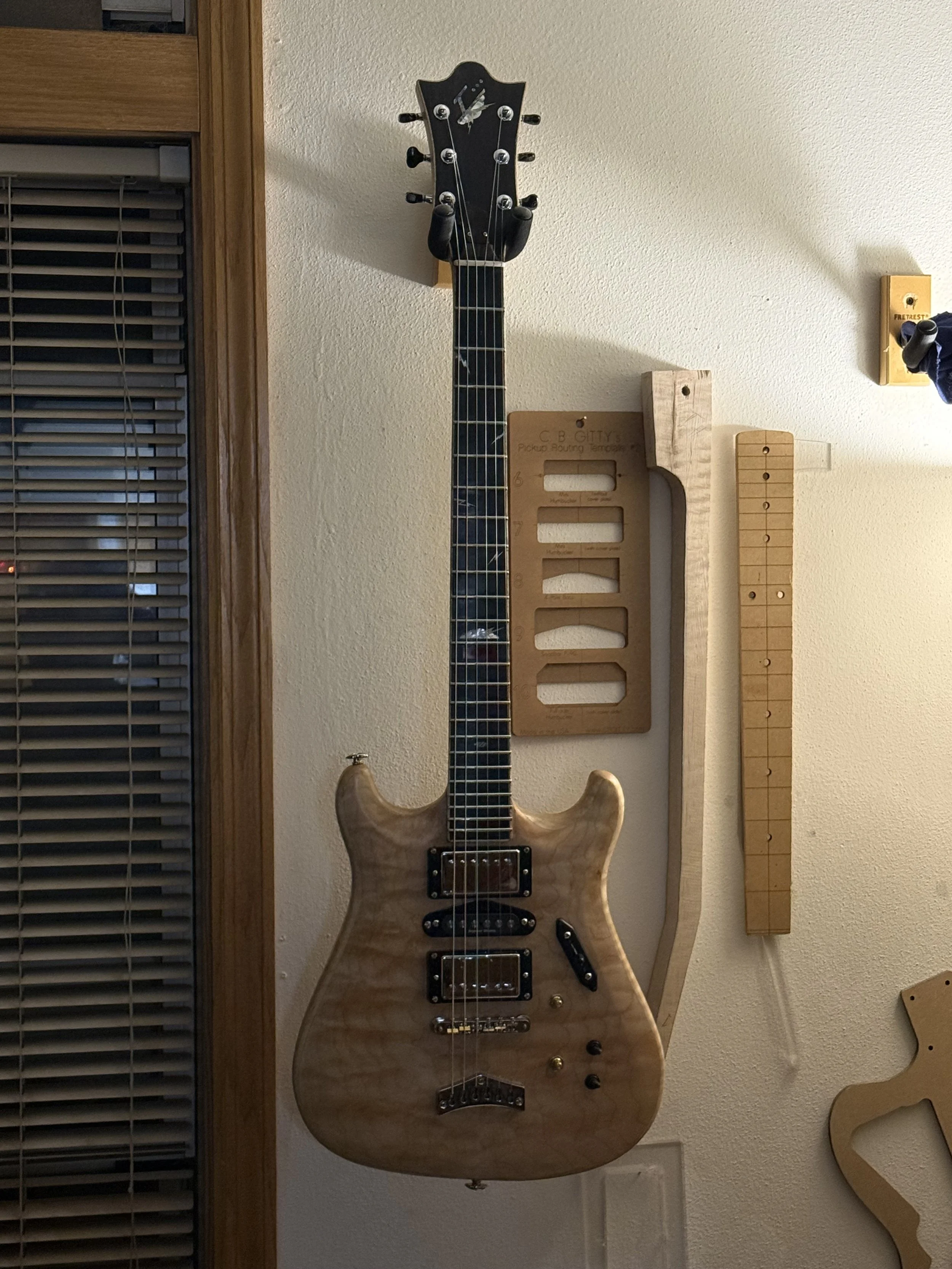 An electric guitar with a natural wood finish hanging on a wall. To the right, there are wooden tools or parts mounted on the wall, and to the left, a window with blinds.