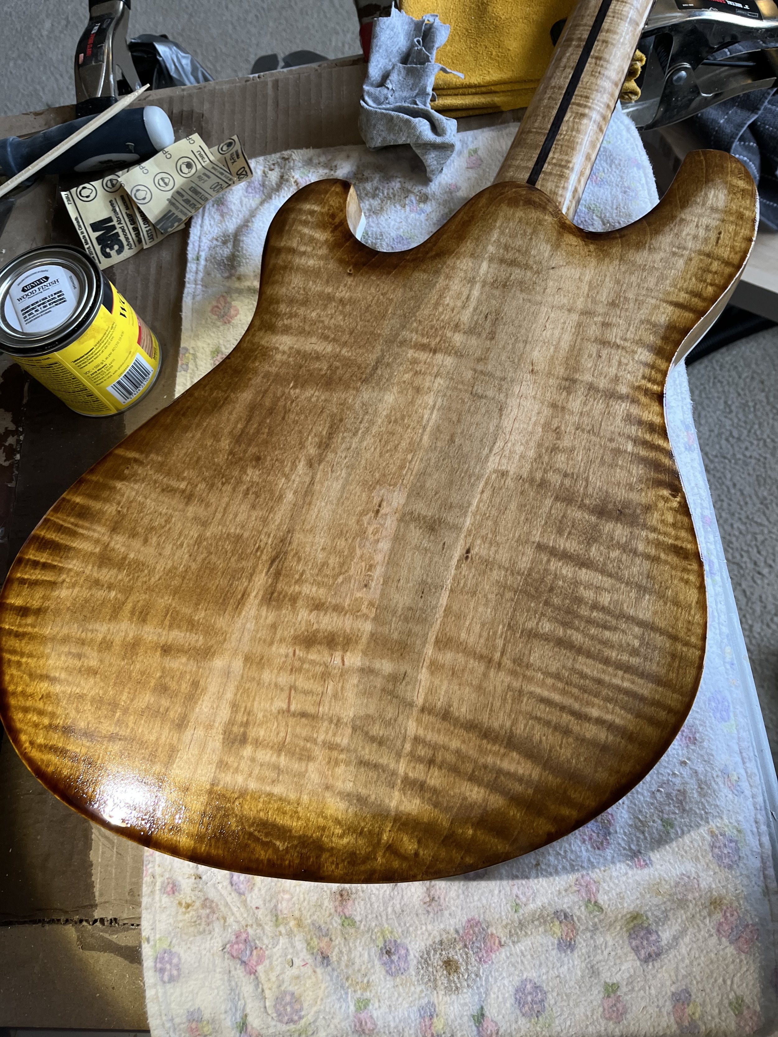 Close-up of a wooden guitar body with a shiny finish, placed on a cloth-covered surface with tools and supplies nearby.