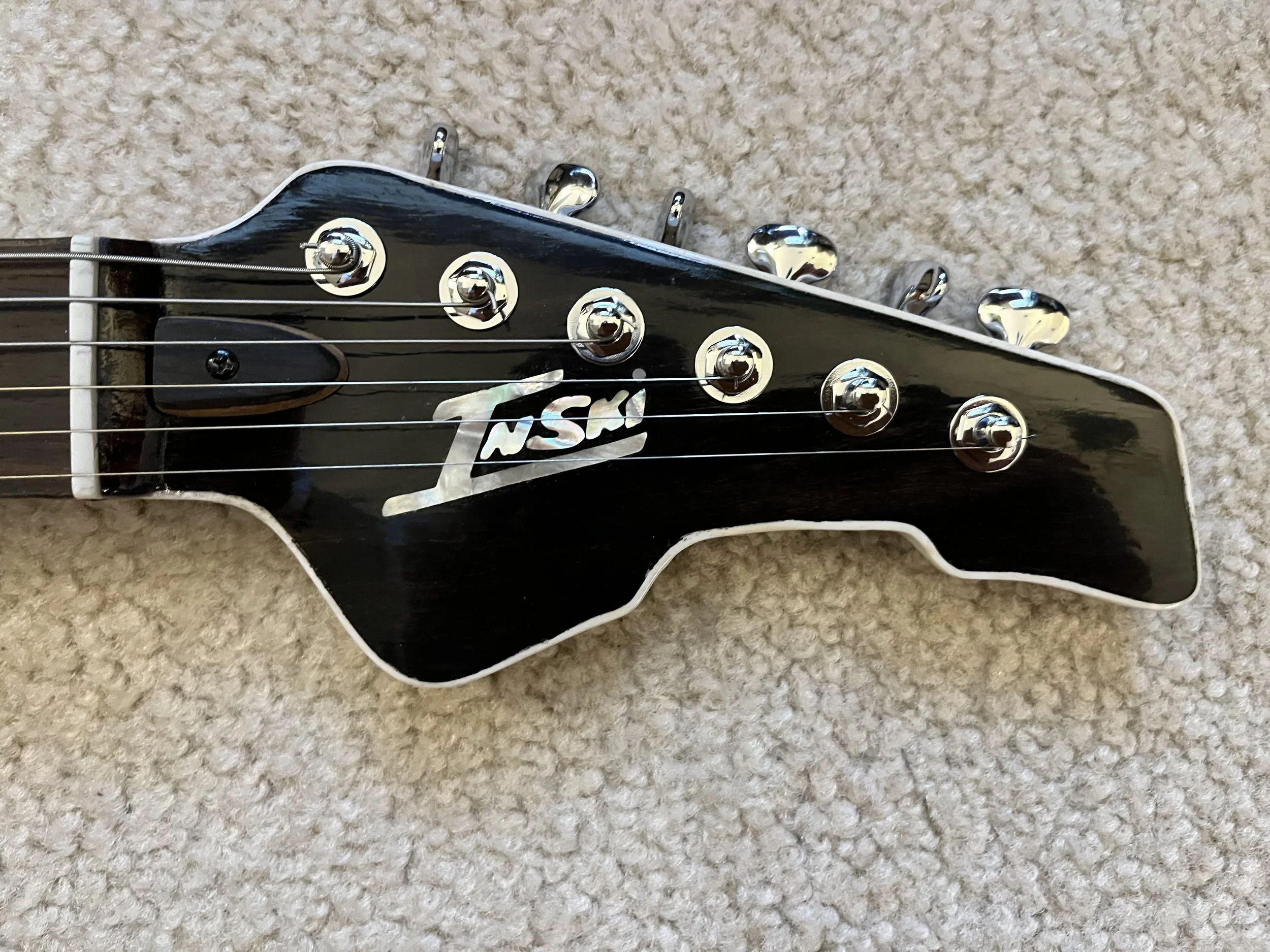 Close-up of a black guitar headstock with six chrome tuning pegs and a white logo on the front, lying on a beige carpet.