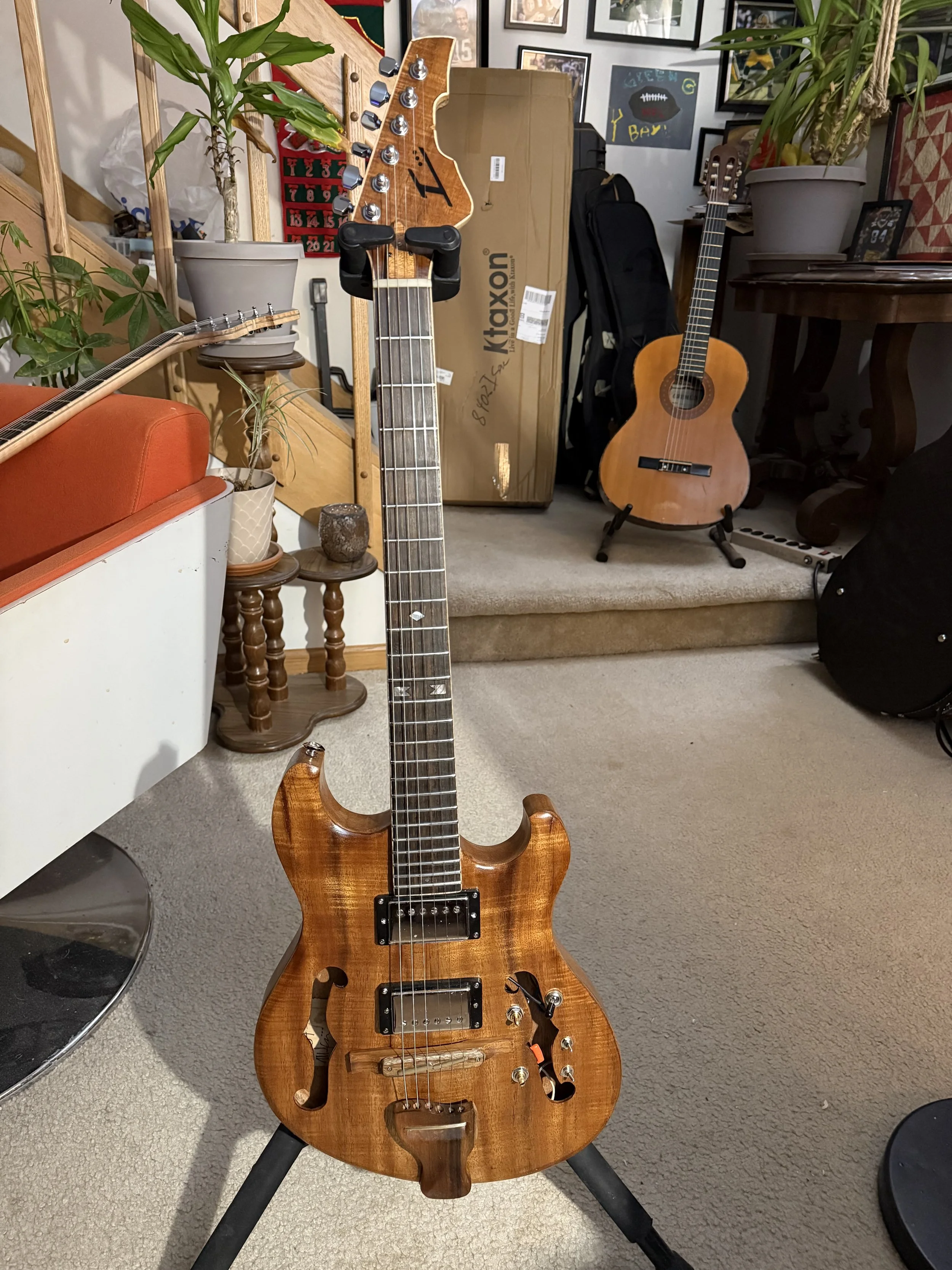 A wooden electric guitar on a stand in a room with various plants, framed photos, and posters on the wall. There is an acoustic guitar leaning against the wall in the background, and a box labeled 'Kitaxon' behind it.