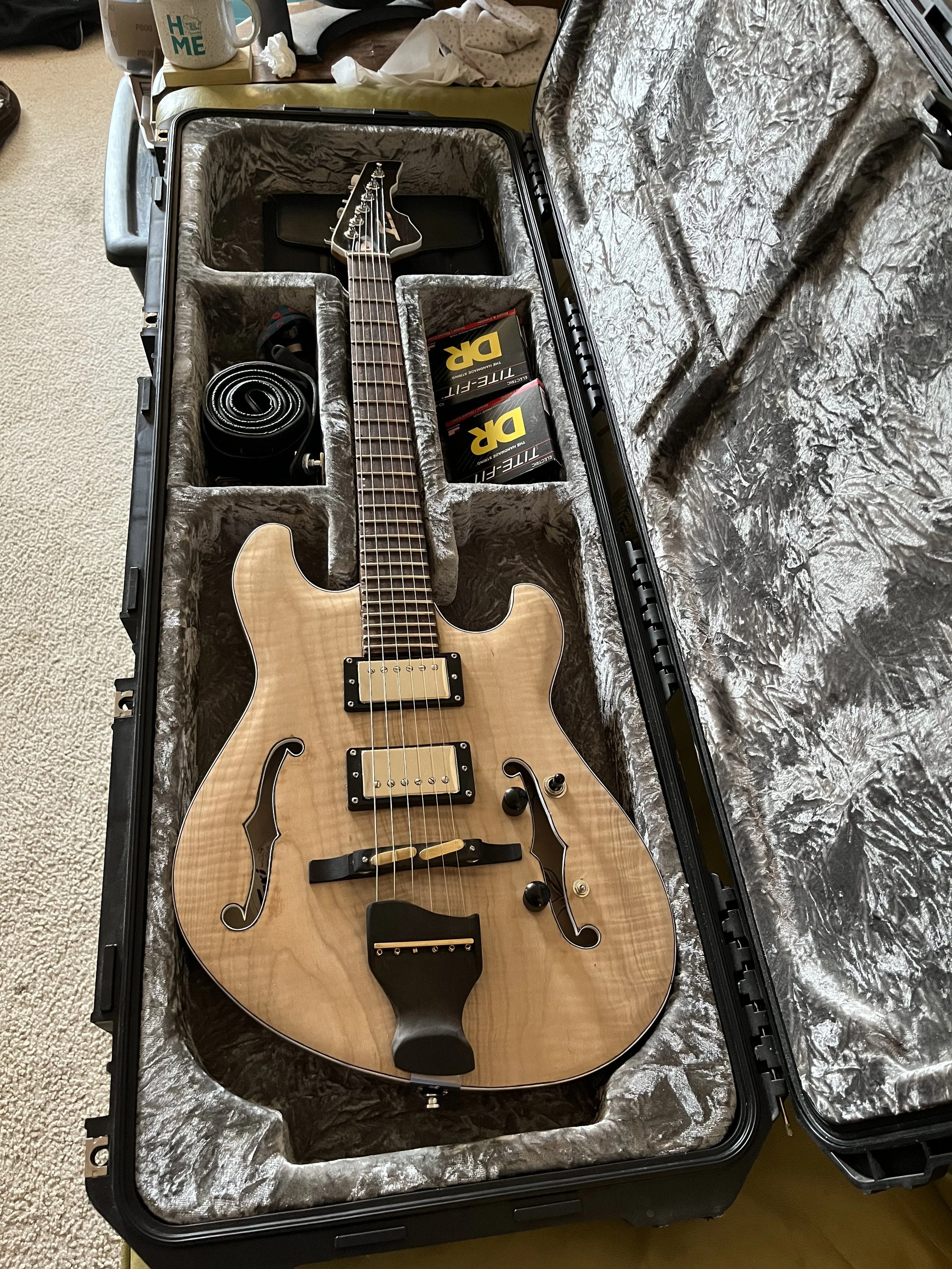 A semi-hollow blonde electric guitar inside a hard case with gray plush lining. The case has compartments holding a coiled black cable and two boxes labeled "D'Addario TITANIUM-FI".