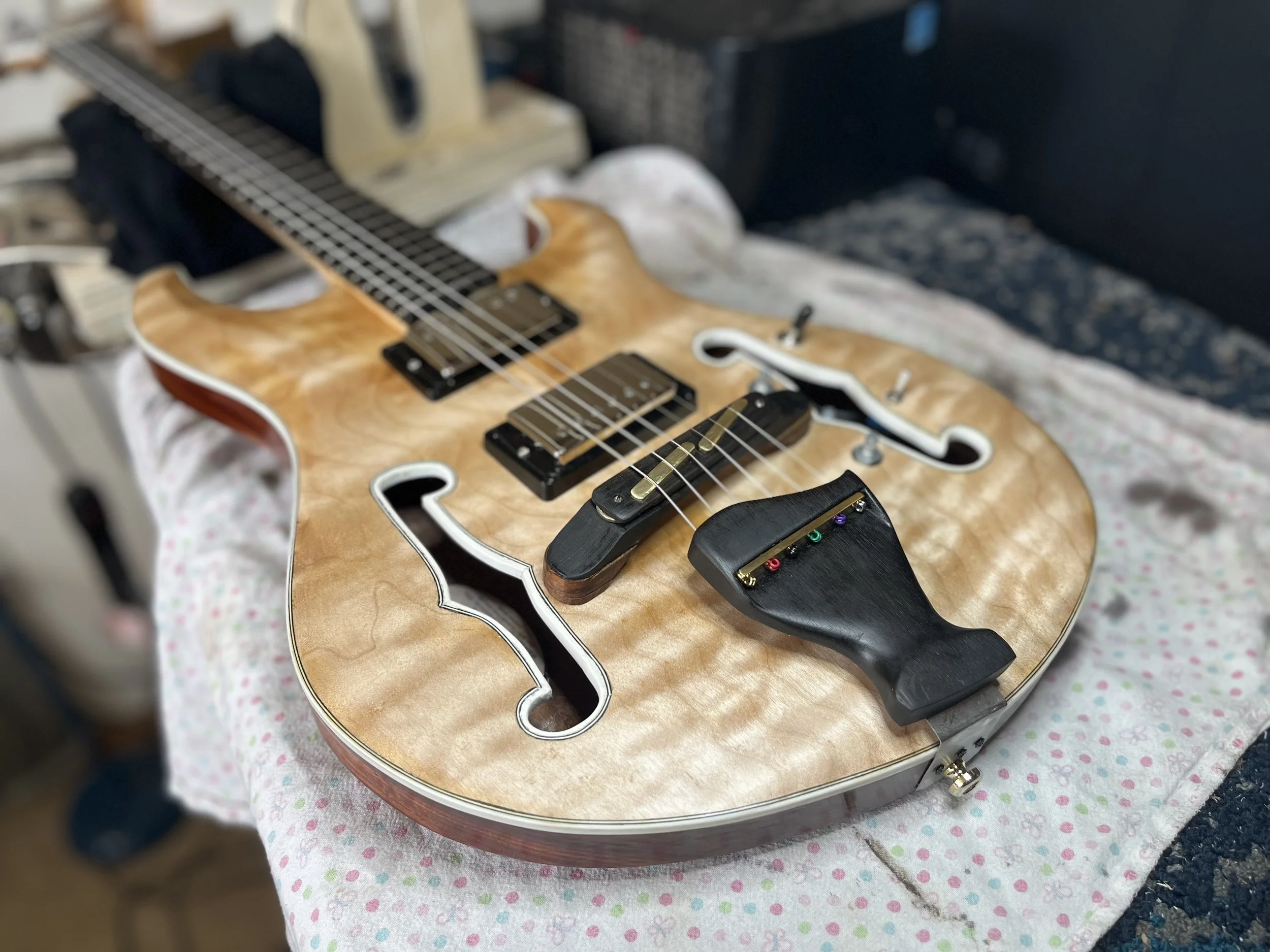 Wooden semi-hollow electric guitar with f-holes and humbucking pickups resting on a patterned cloth. The guitar has a tortoiseshell pickguard and gold hardware. In the background, there is a black amplifier and other electronic devices.