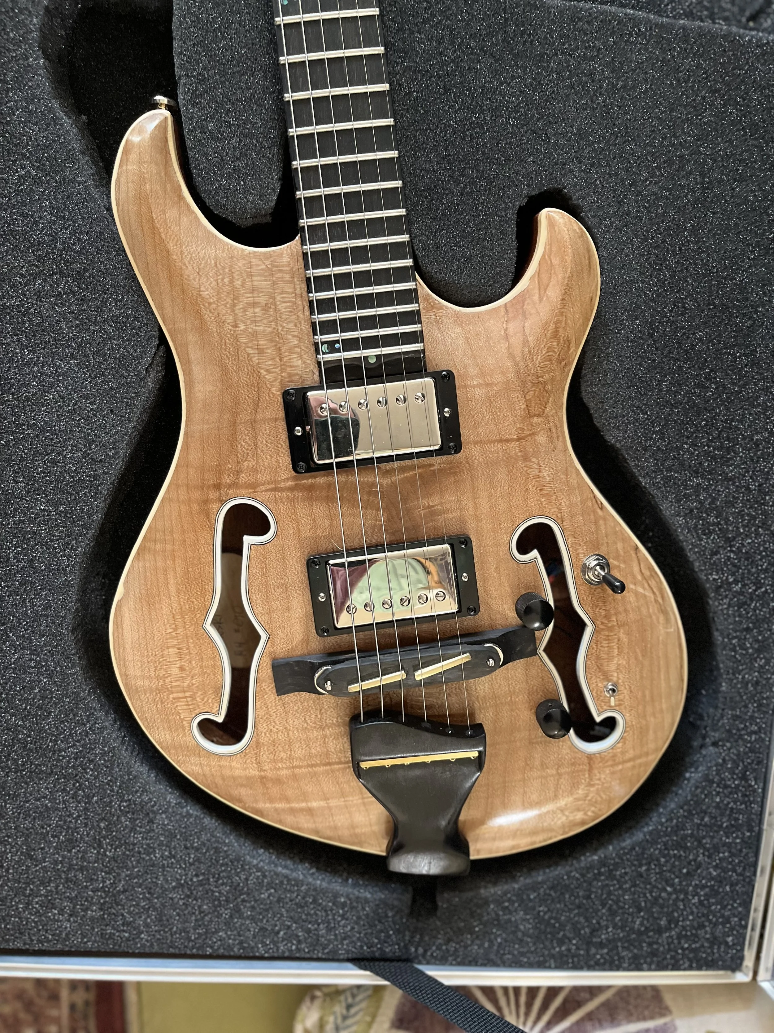 A semi-hollow electric guitar with a natural wood finish in a black case with foam padding.