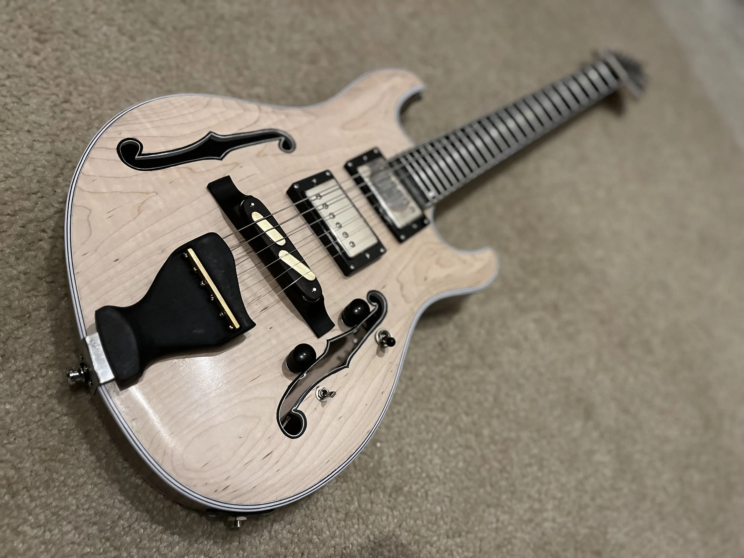 A semi-hollow electric guitar with a natural wood finish, black pickguard, and F-style body shape, resting on a beige carpet.
