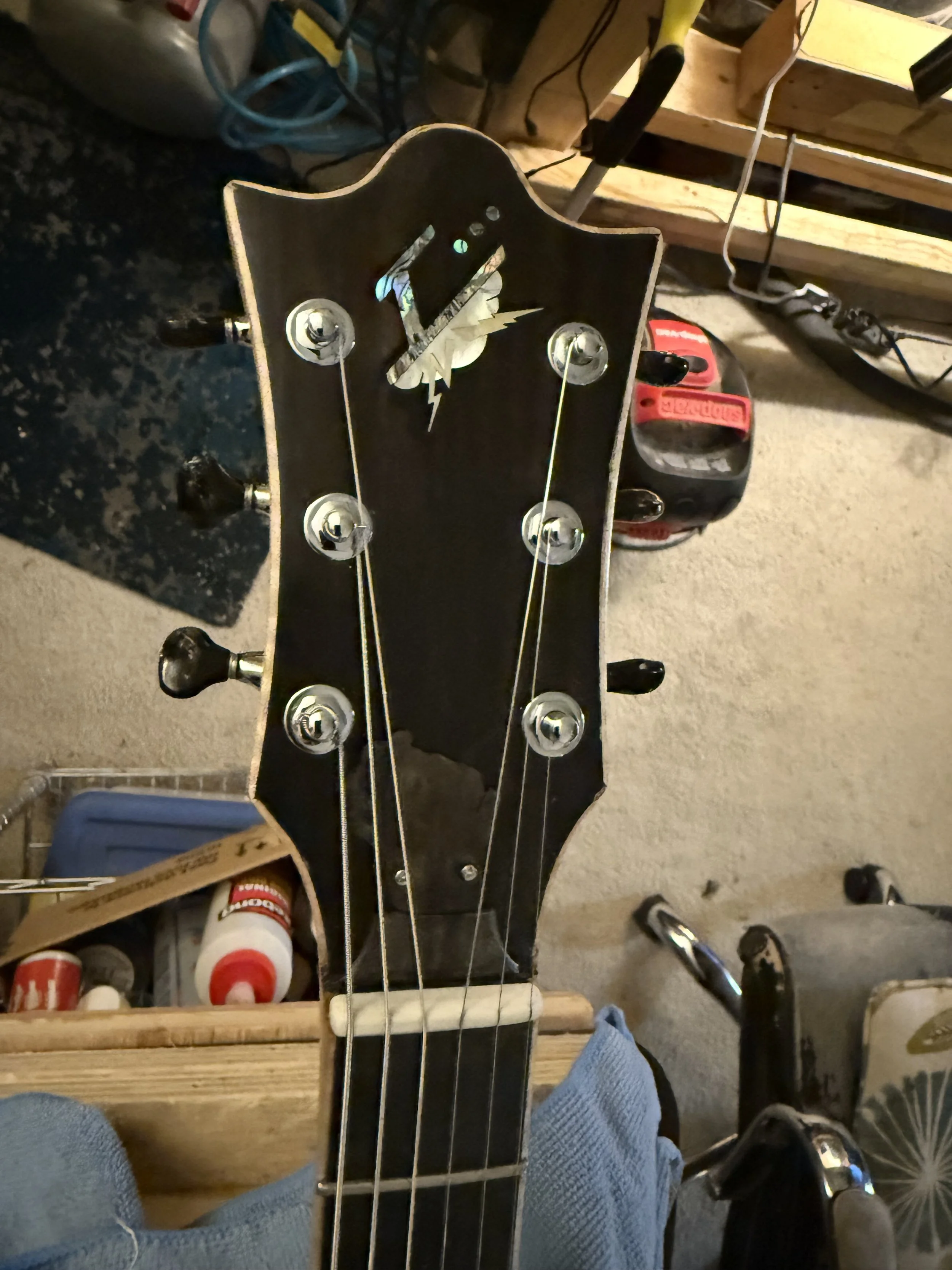 Top view of a black guitar headstock with tuning pegs, against a cluttered workshop background with tools and supplies.